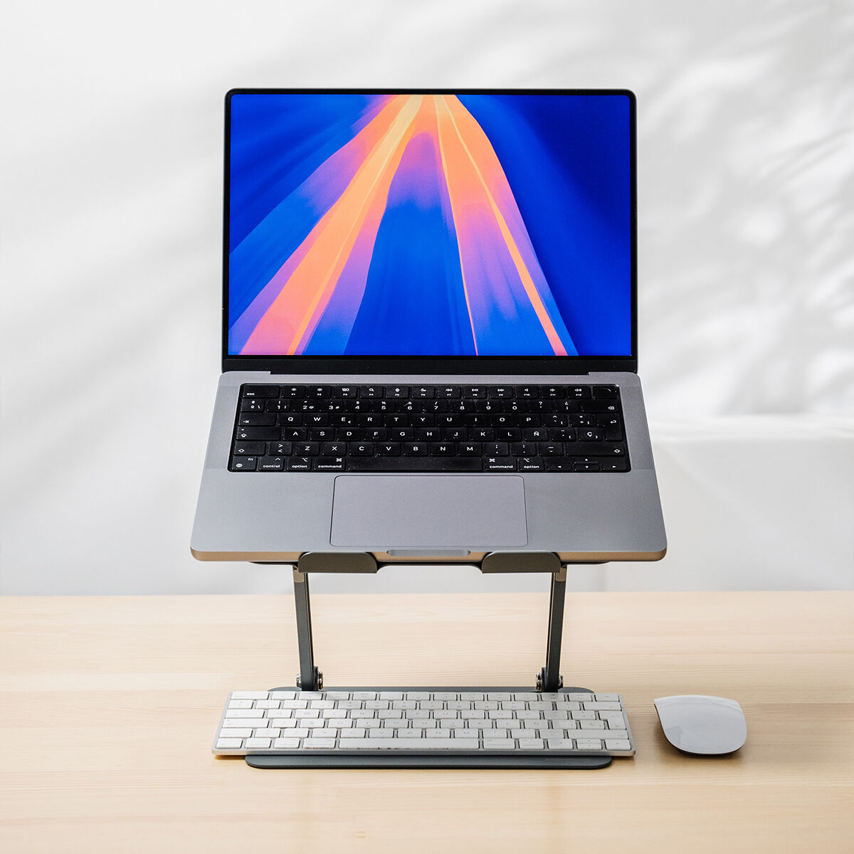 Laptop argento su supporto, con tastiera e mouse su tavolo in legno chiaro.