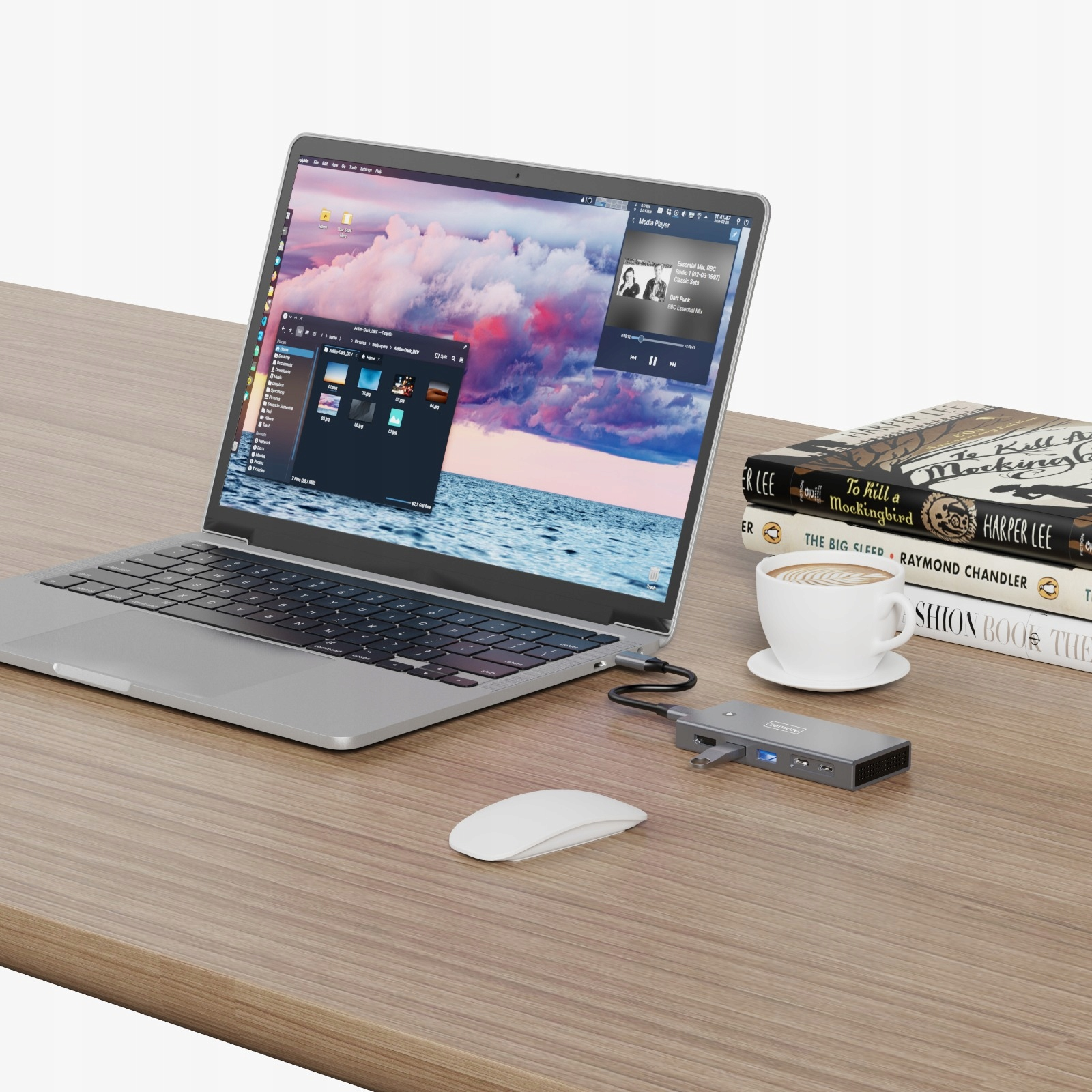 Laptop na drewnianym biurku z myszą, zewnętrznym hubem, filiżanką kawy i książkami. Ekran pokazuje muzykę.