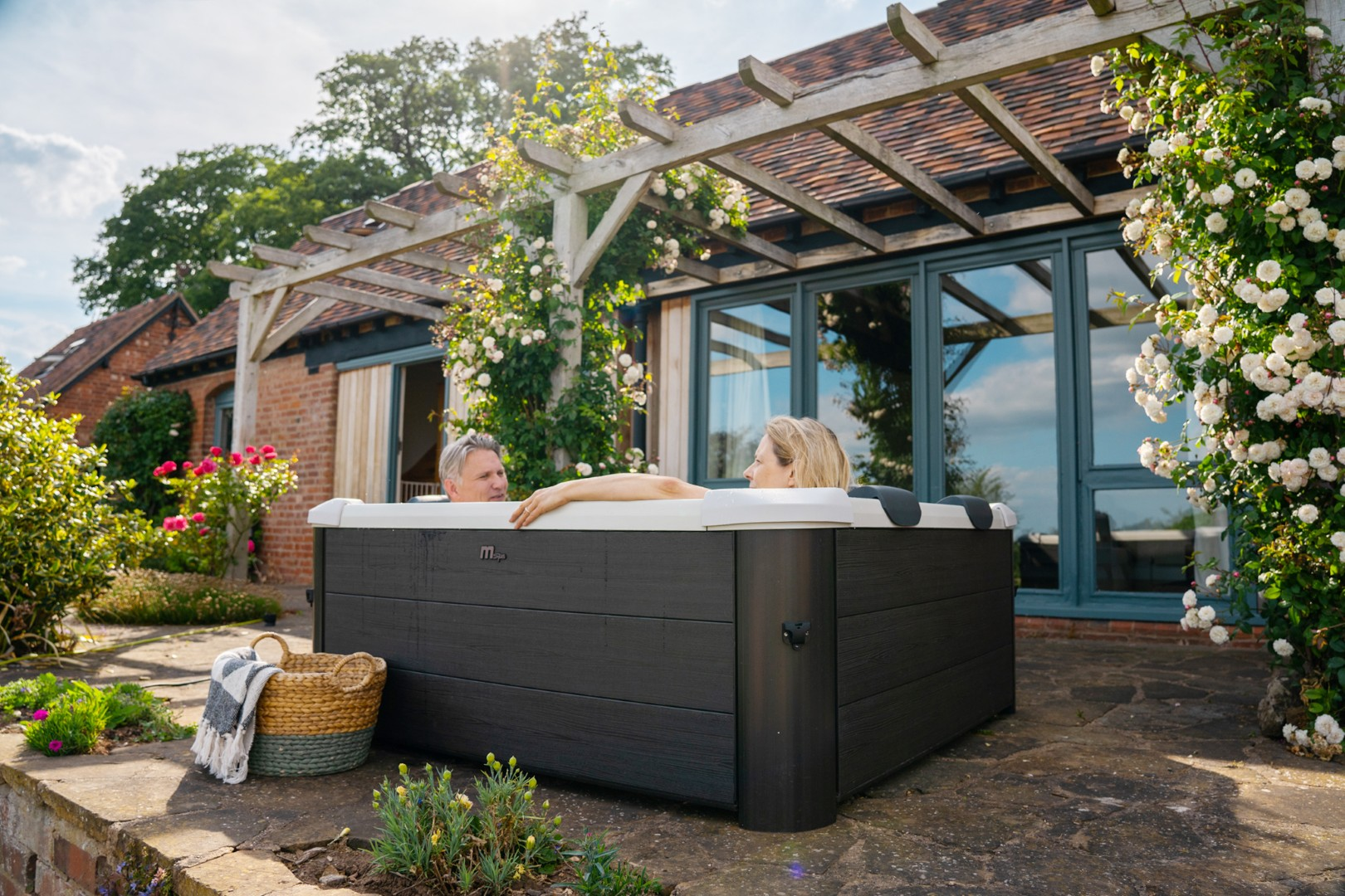Ein Paar entspannt in einem dunklen Whirlpool in einem Garten mit Pergola und Backsteinhaus.