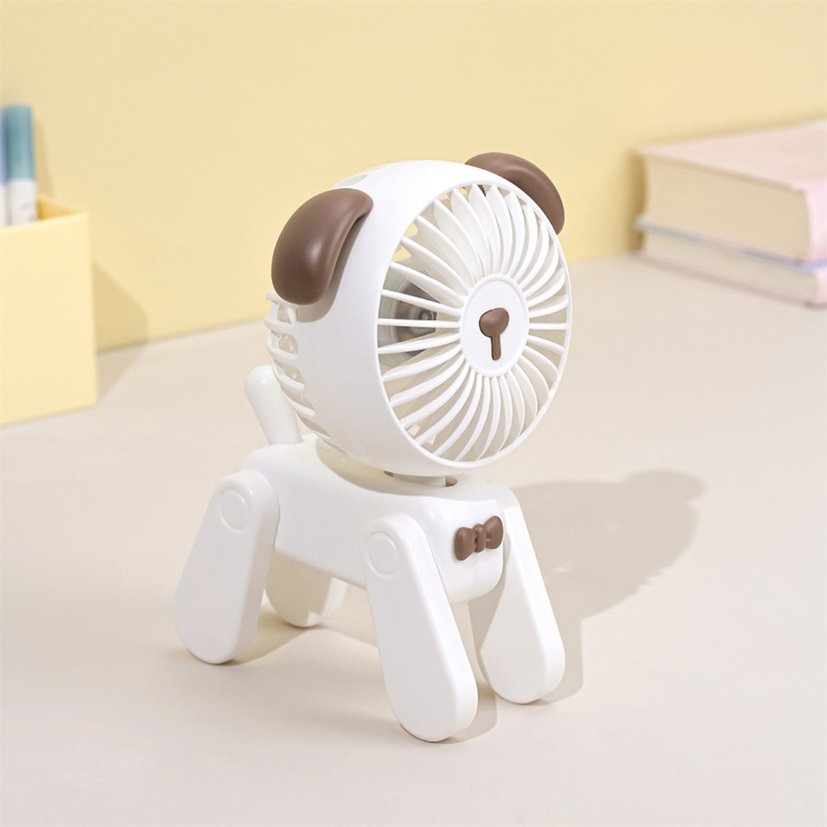 Weißer hundeförmiger Ventilator auf einem Schreibtisch. Es hat braune Ohren und eine Fliege. Der Hintergrund ist eine hellgelbe Wand.