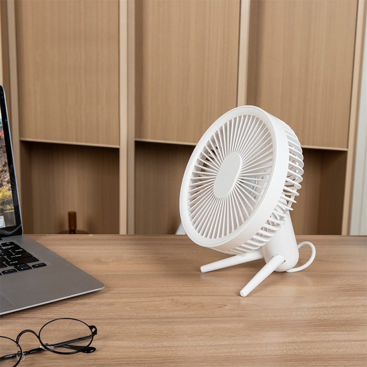 Weißer Schreibtischventilator auf einem Holztisch. Ein Laptop, eine Brille und ein Holzschrank im Hintergrund.
