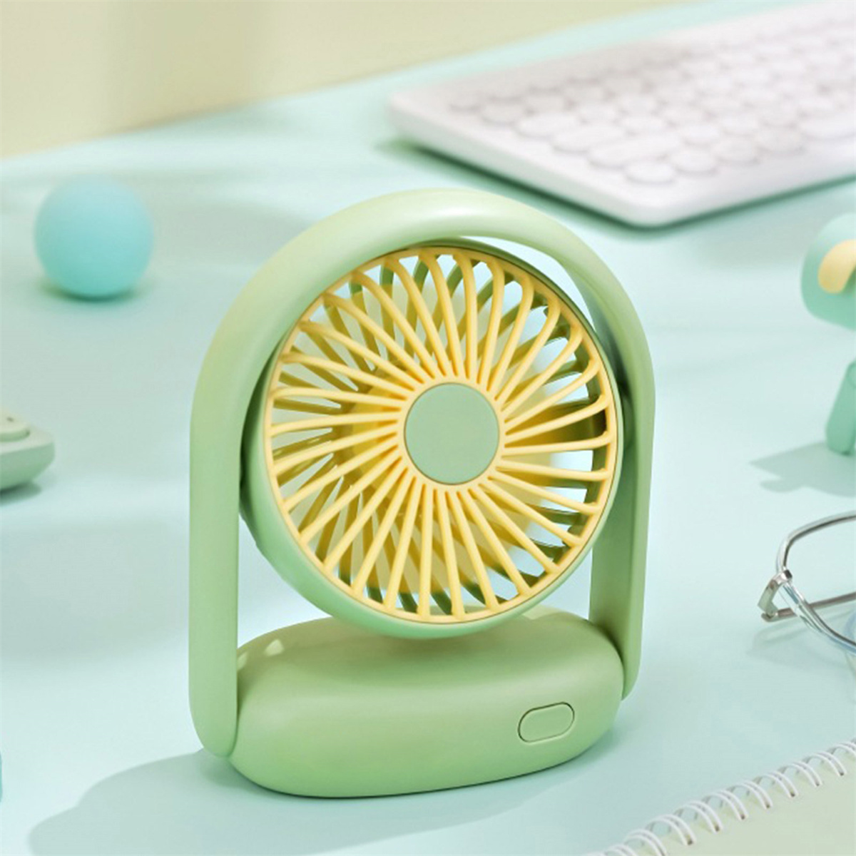 Ein kleiner grüner Ventilator auf einem Schreibtisch. Er hat ein gelbes Lüfterrad und einen Griff.