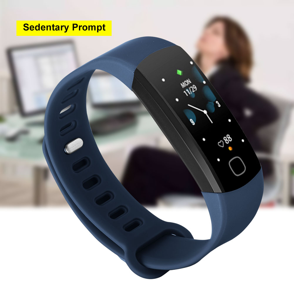 Blauer Fitness-Tracker, mit Bildschirm, der Zeit und Herzfrequenz anzeigt. Im Hintergrund sitzt eine Frau am Schreibtisch.