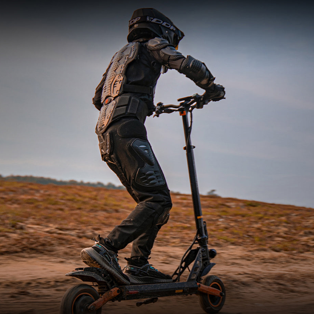Persona montando un patinete eléctrico negro y naranja en un camino de tierra.