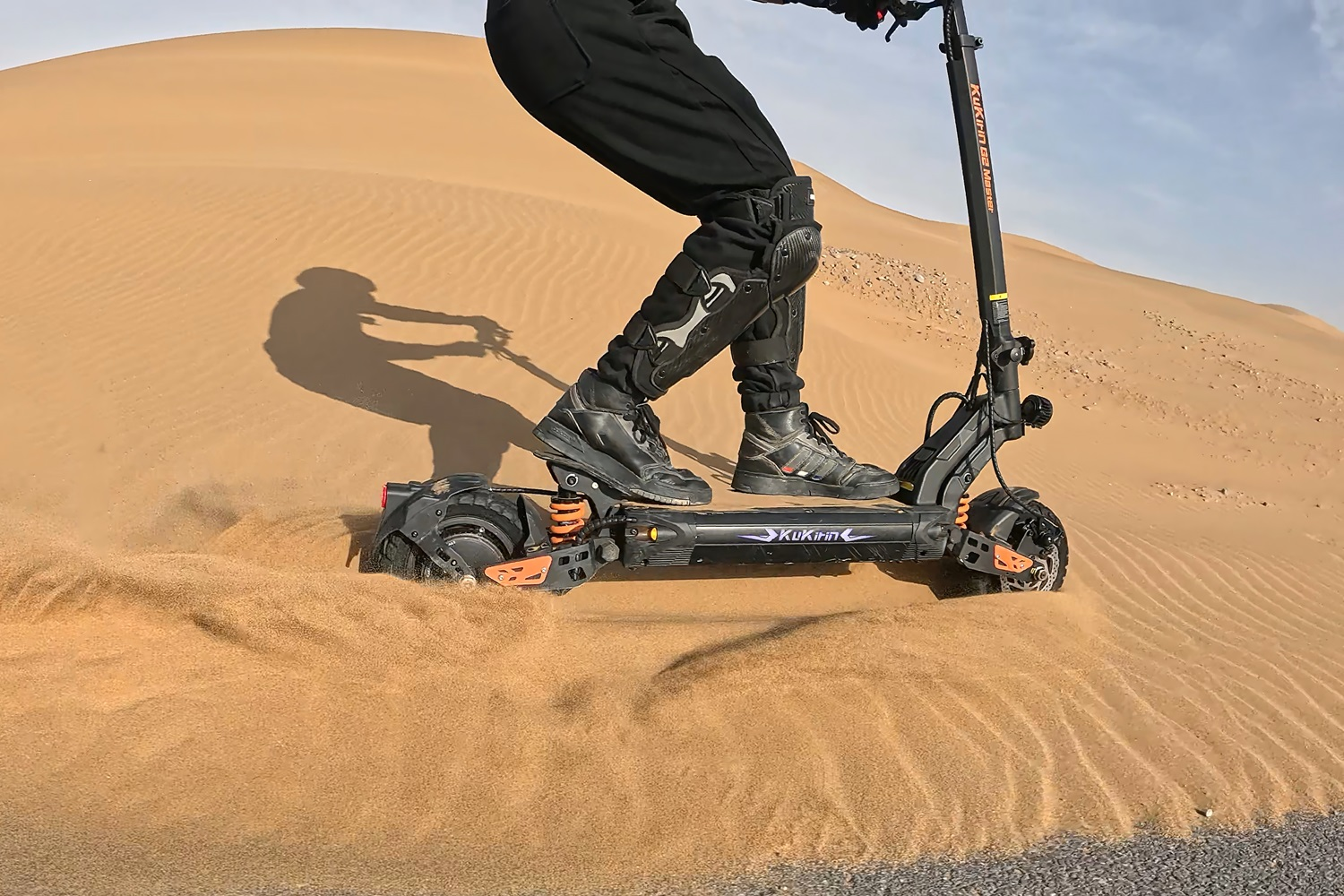 Una persona su un monopattino elettrico nella sabbia del deserto. Lo scooter è nero e arancione.