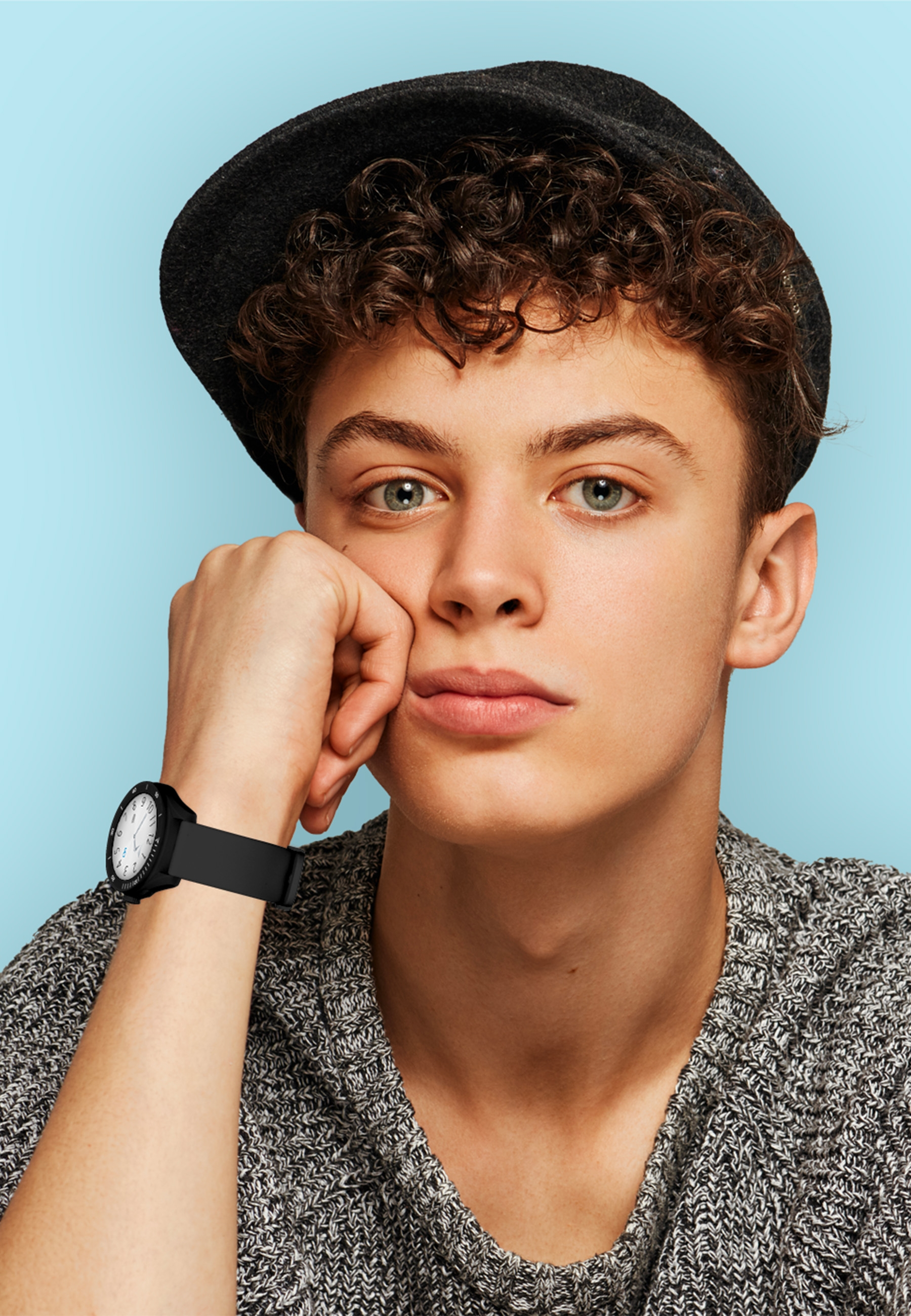 Jeune homme portant un chapeau noir et une montre, reposant son menton sur sa main, fond bleu.