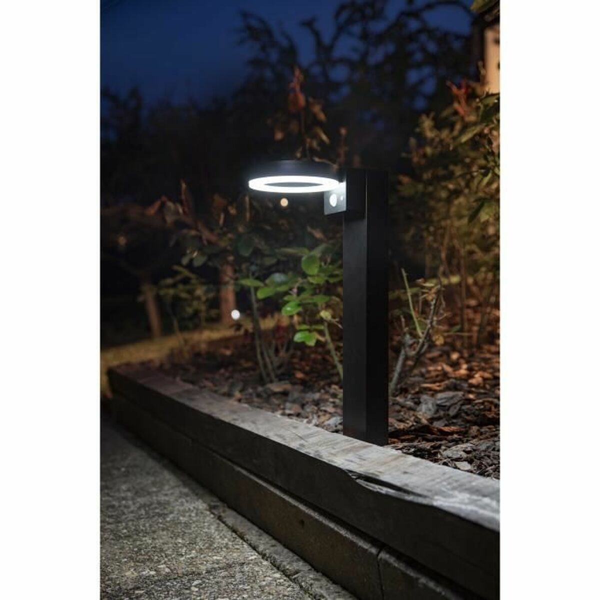 Schwarze Außenleuchte. Die Lampe hat ein Ringlicht oben. Hintergrund: Garten und Bäume.