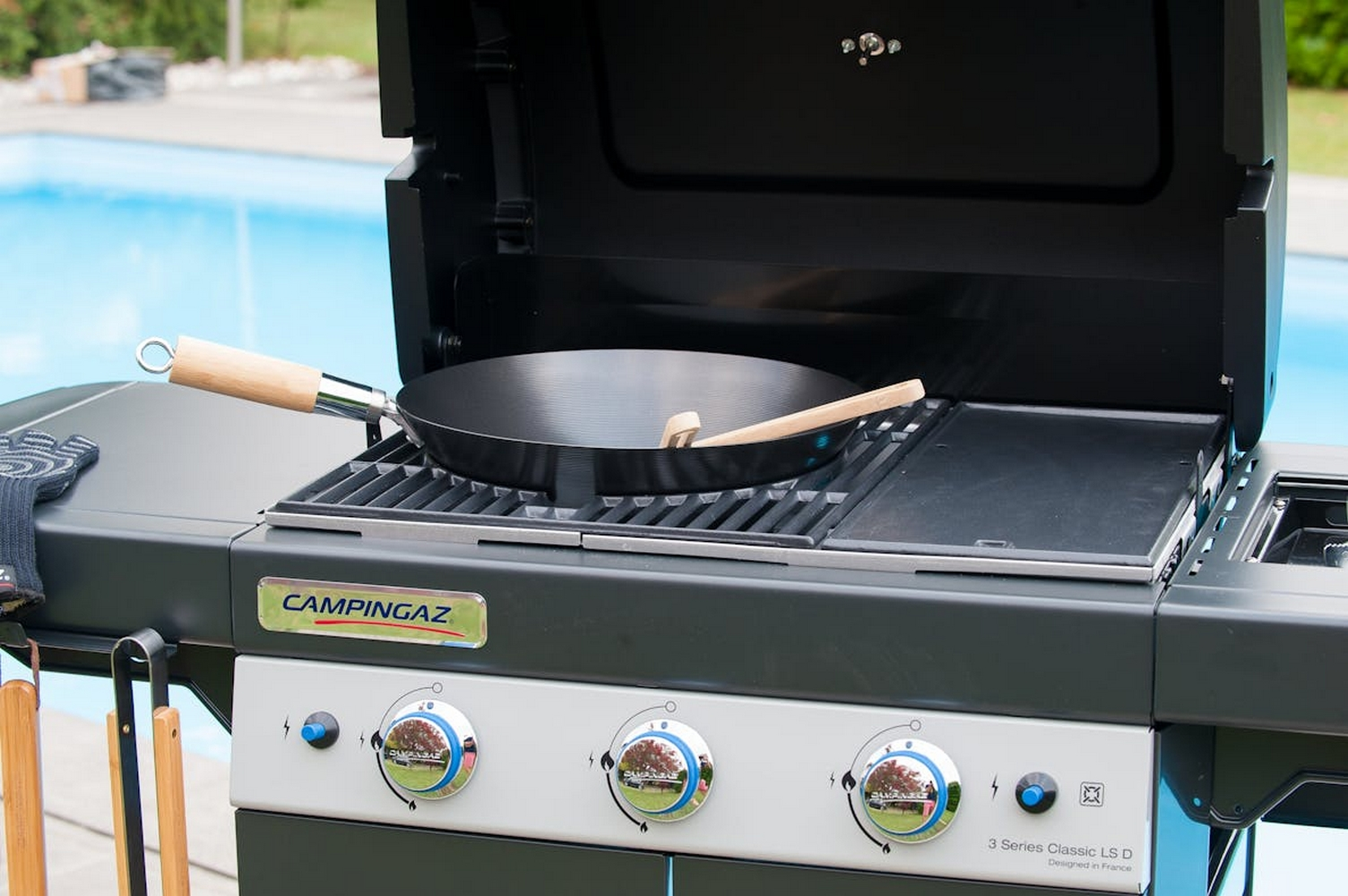 Un barbecue nero con un wok e un cucchiaio di legno sopra, e una piscina sullo sfondo.
