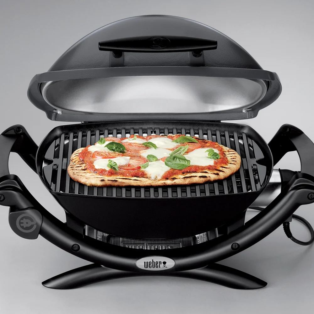Una parrilla Weber negra con tapa abierta sostiene una pizza con albahaca y tomates.