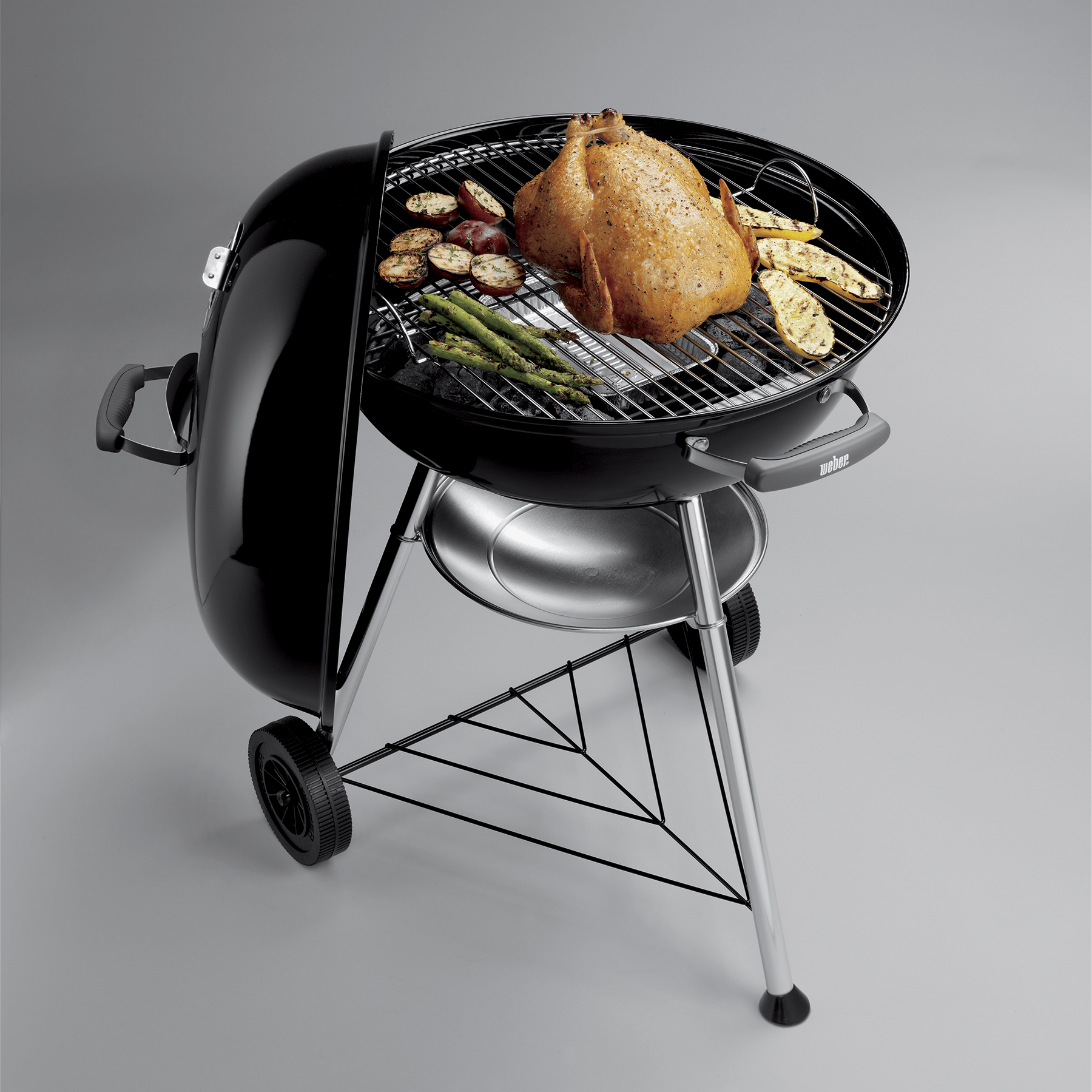 Griglia Weber nera con coperchio aperto, mostra pollo alla griglia, verdure e carbone.