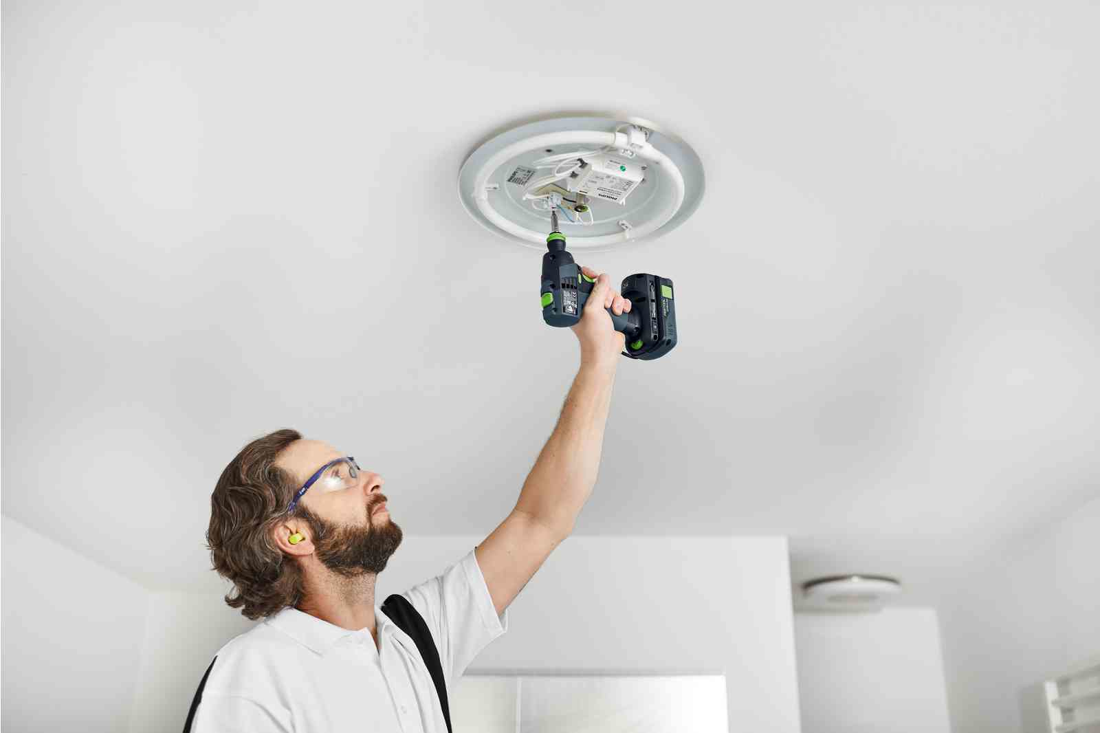 Mężczyzna w białej koszuli używa wiertarki do montażu lampy sufitowej. Nosi okulary ochronne i zatyczki do uszu.