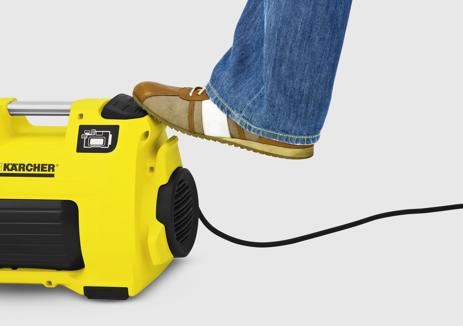 Żółta myjka ciśnieniowa Kärcher z nogą na niej. Widoczne dżinsy i brązowy but. Białe tło.