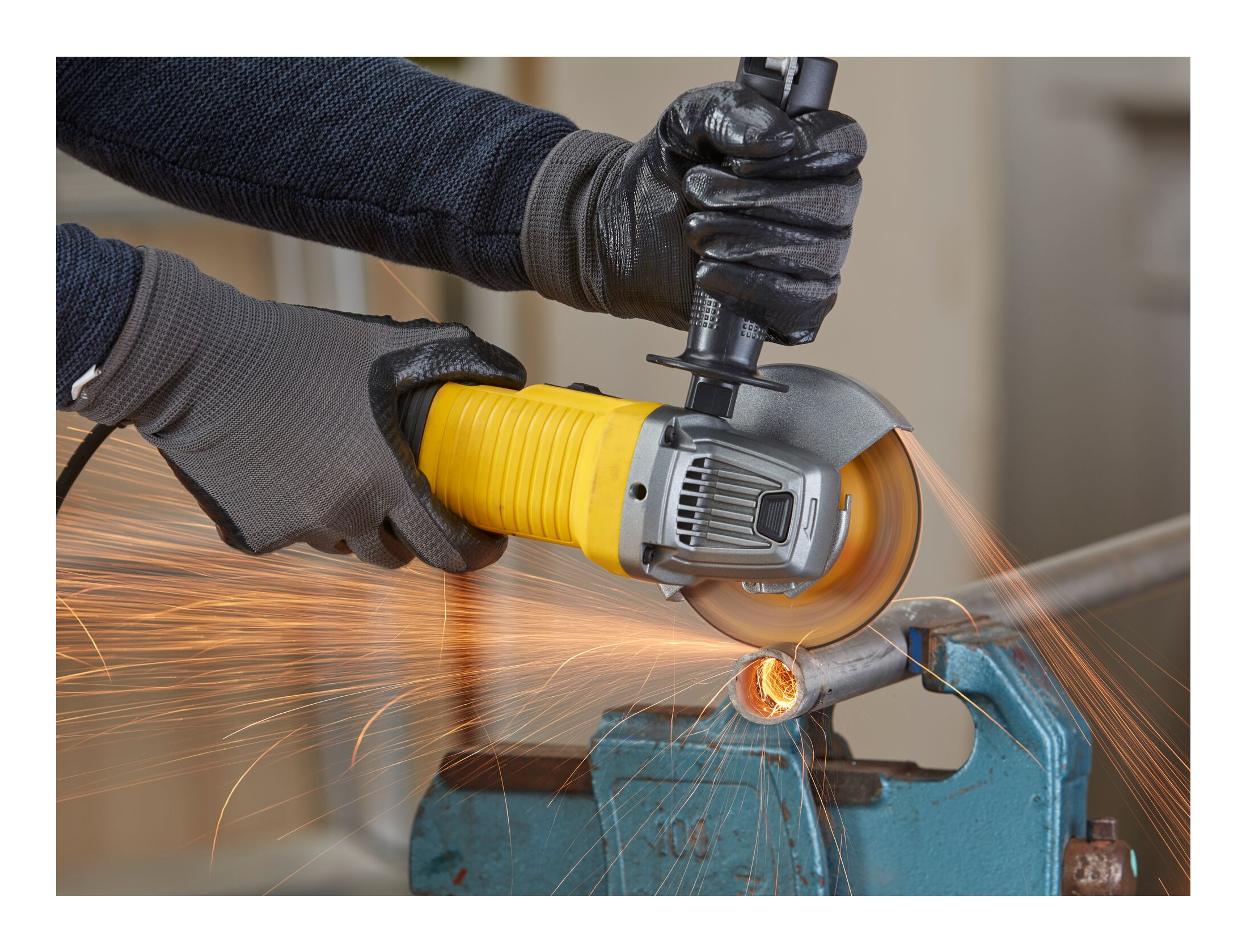 Robotnik tnie metalową rurę żółtą szlifierką kątową. Iskry lecą. Noszone są czarne rękawice.