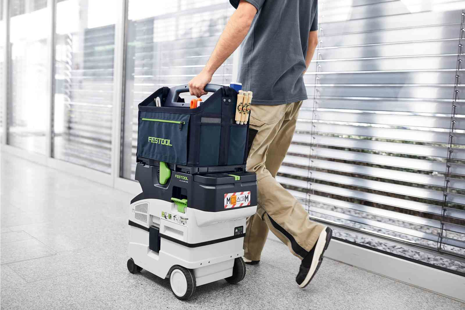 Ein Mann trägt eine Werkzeugkiste und einen Trolley mit Werkzeug auf weißem Boden.