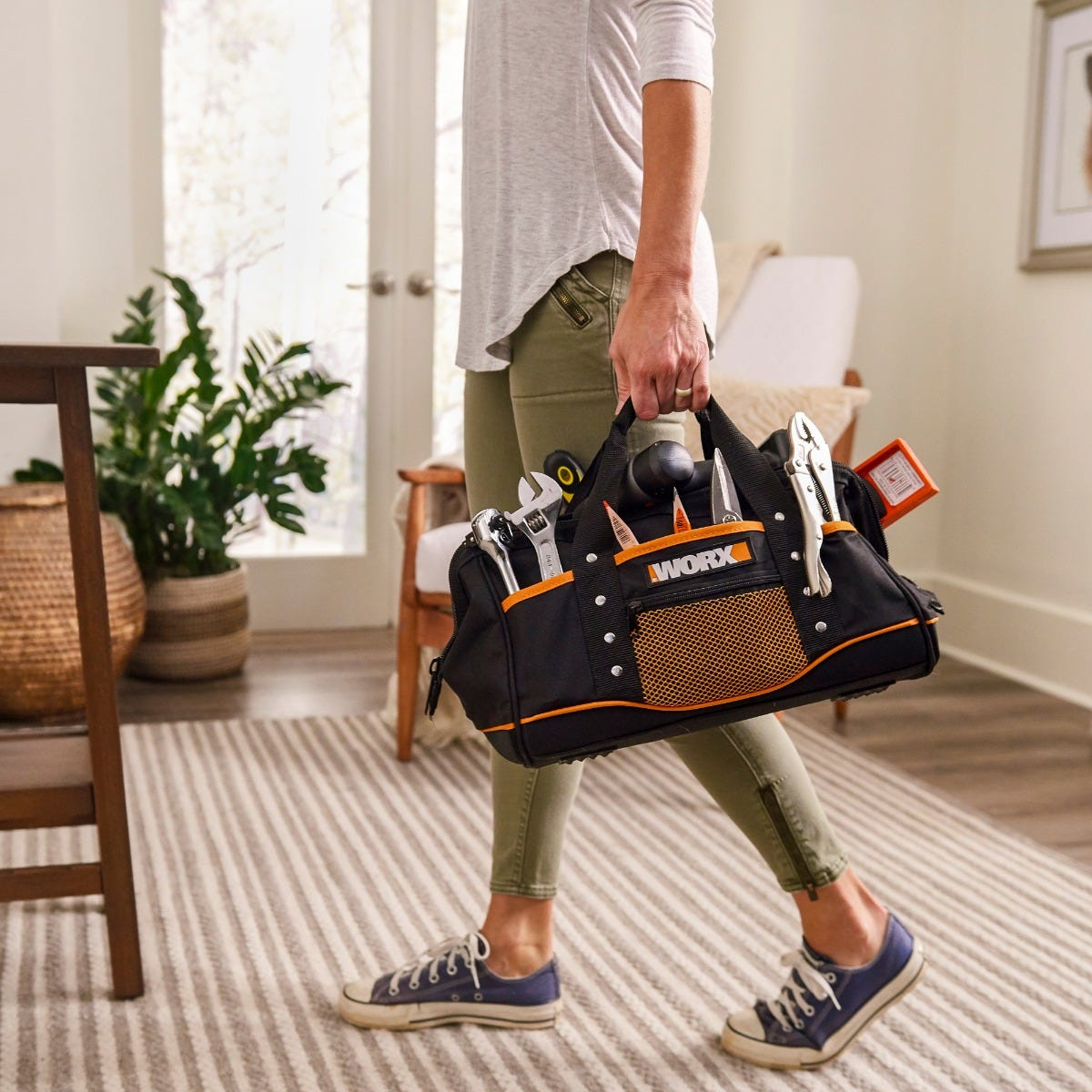 Frau hält eine schwarze und orangefarbene Werkzeugtasche mit Werkzeugen in einem häuslichen Umfeld.