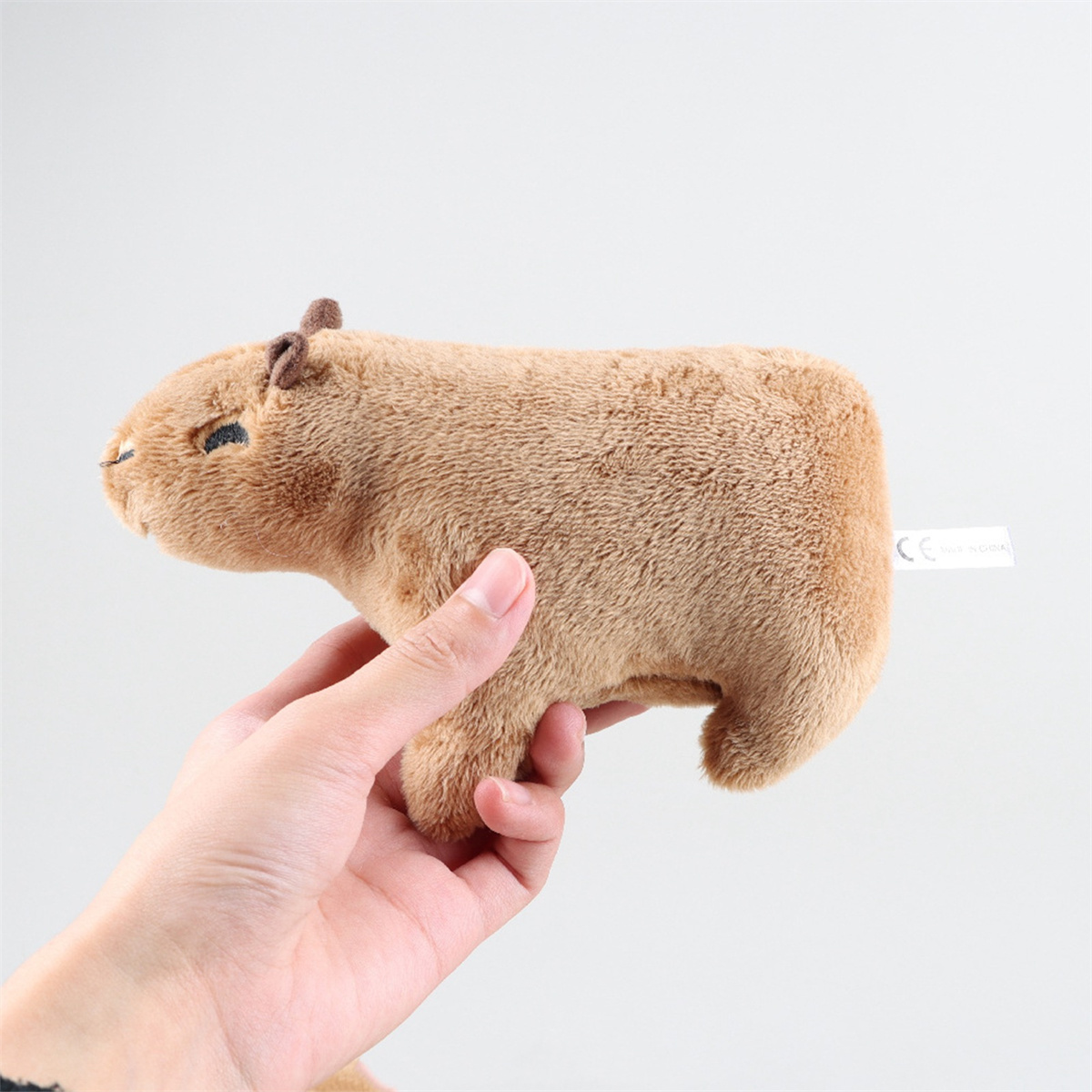 Hand einer Person, die ein braunes Capybara-Plüschtier hält. Weißer Hintergrund.