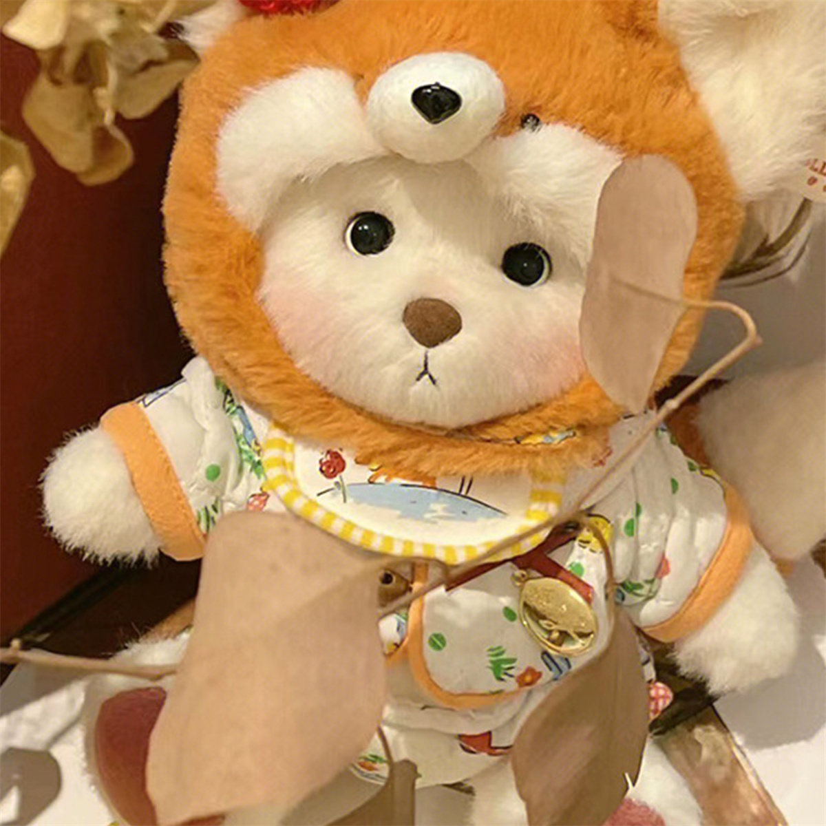 Teddybär mit orangefarbener Bärenkapuze und gemustertem Outfit. Braunes Fell, kleine Augen sichtbar.
