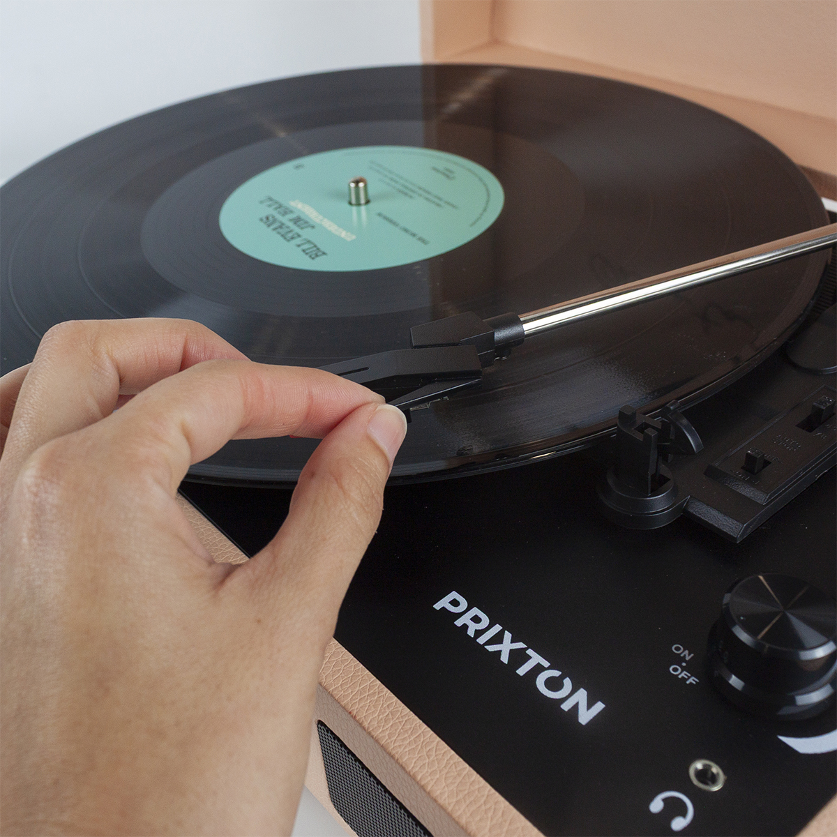 Ręka umieszcza igłę na czarnej płycie winylowej na różowym gramofonie. Widoczny jest napis 'PRIXTON'.