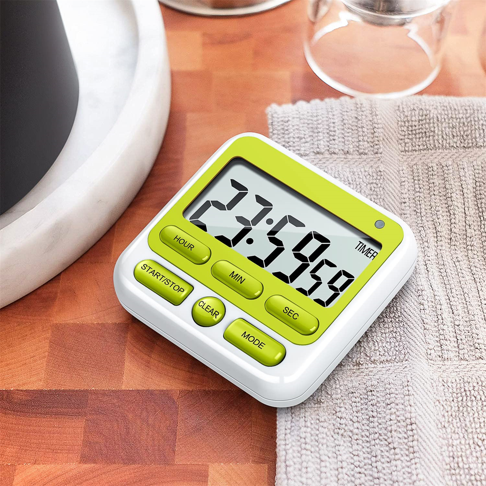 Ein digitaler Küchentimer mit grünen Knöpfen auf einer Holzoberfläche neben einem Handtuch.