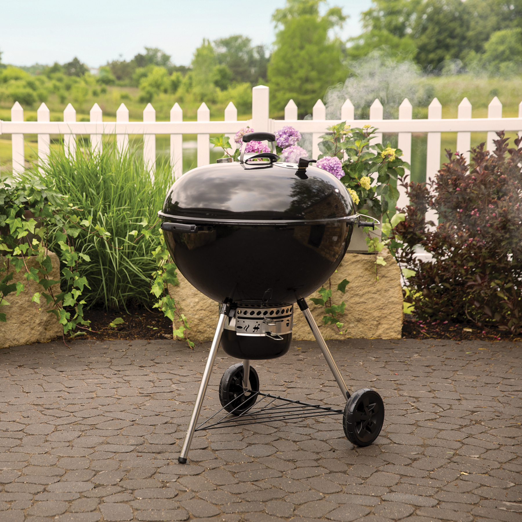 Schwarzer Weber-Grill mit weißem Gartenzaun und Laub im Hintergrund.