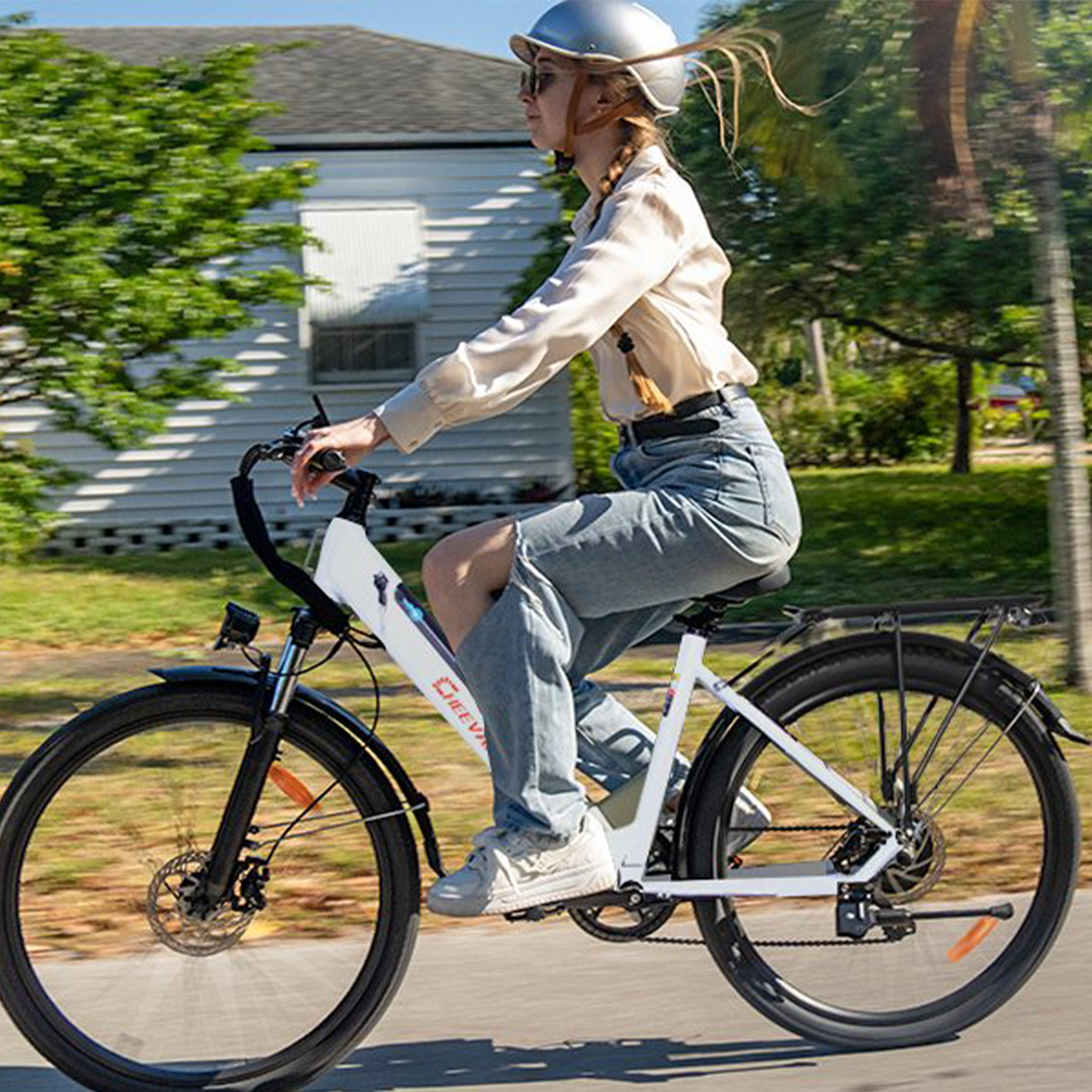 Kobieta jadąca na białym rowerze w słoneczny dzień. Ma na sobie kask, koszulę i dżinsy. Zielone drzewa w tle.