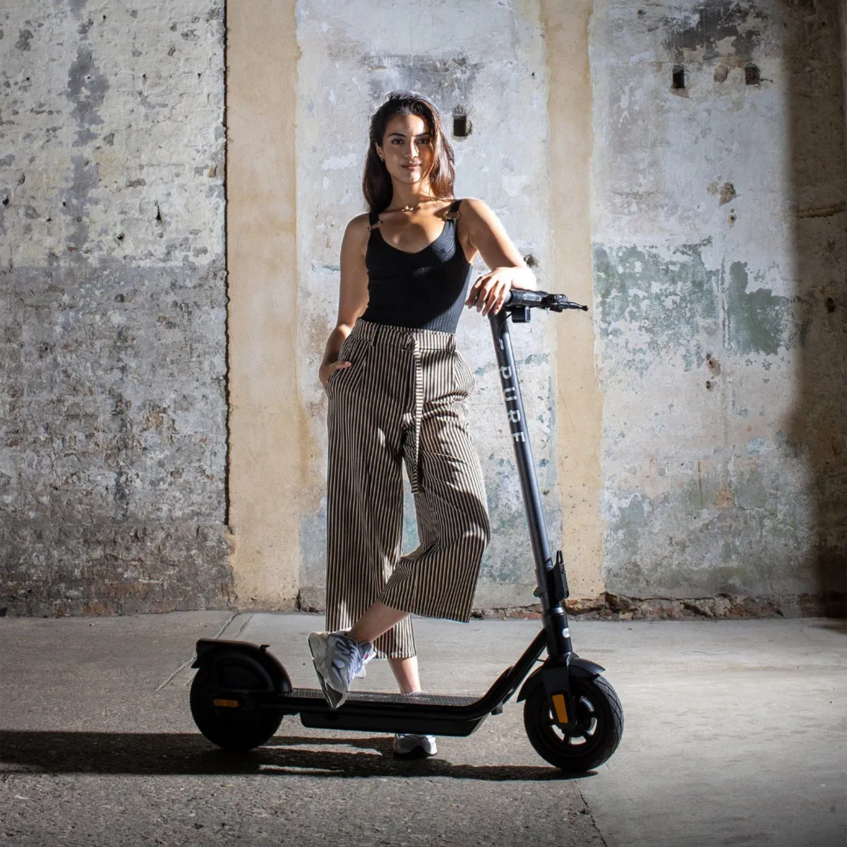 Frau in schwarzem Top und gestreiften Hosen steht mit einem schwarzen Roller in einem schäbigen Raum.