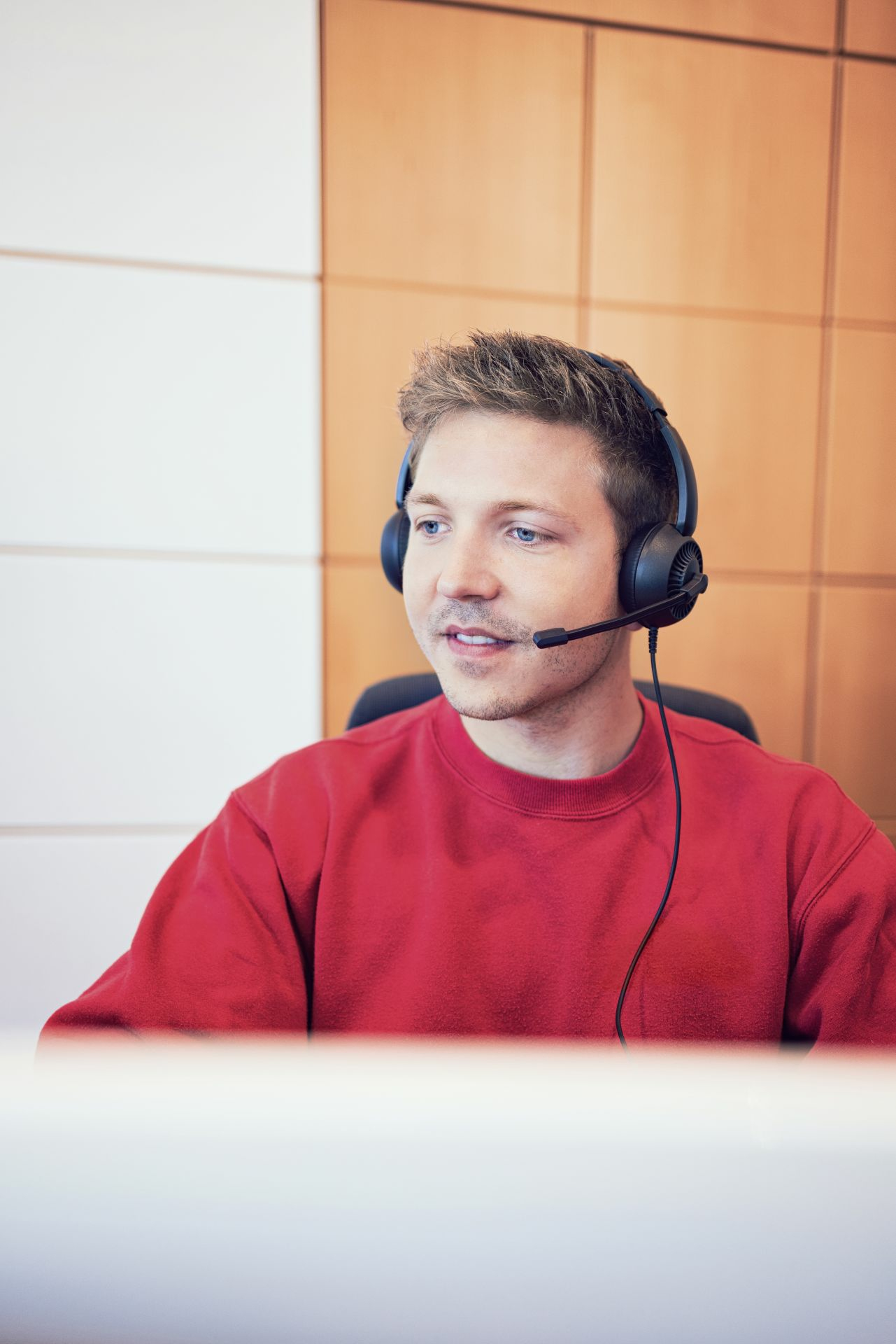 A man wearing a headset, red sweater, sits in front of a wall of white and wood panels.