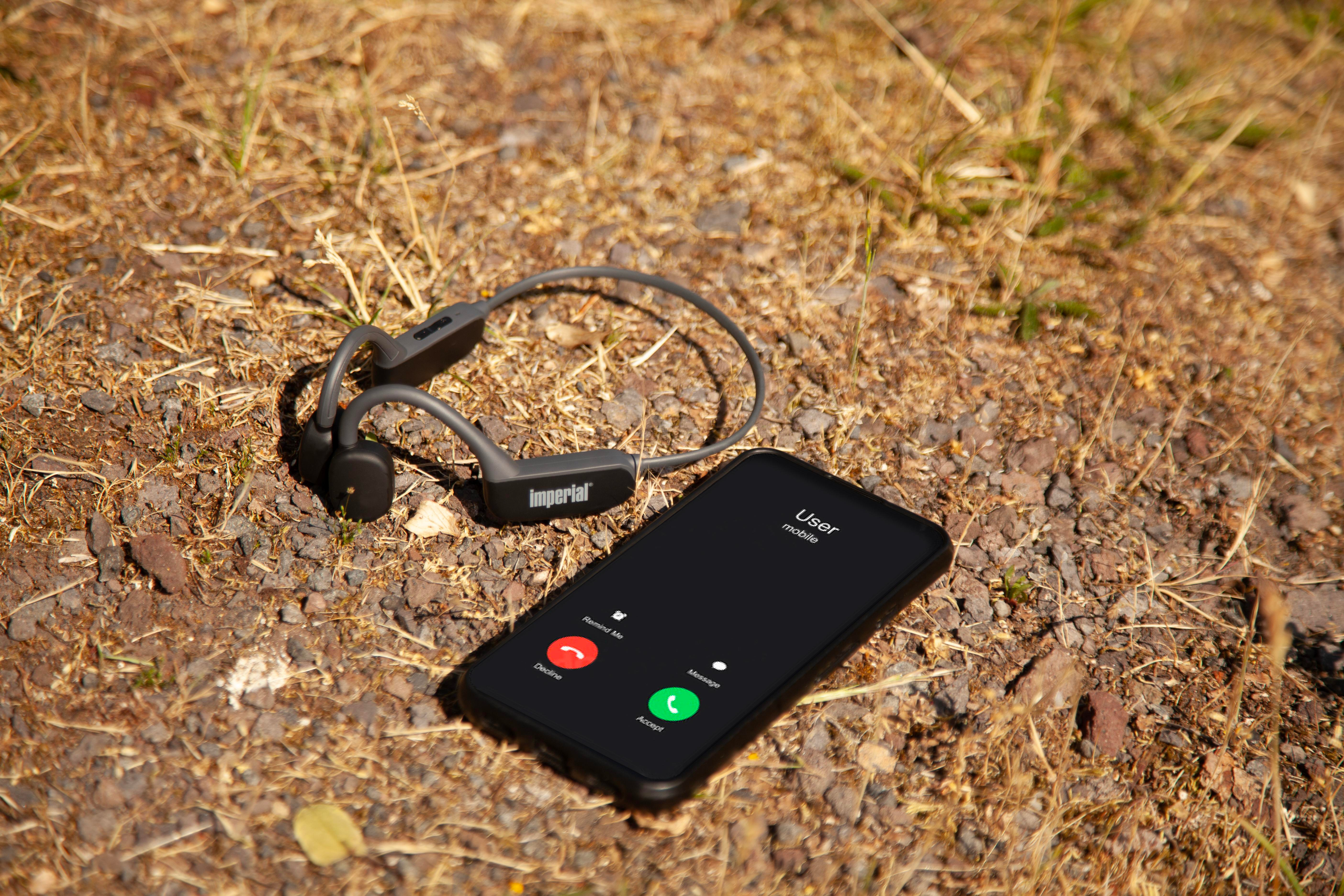 Słuchawki kostne na ziemi ze smartfonem pokazującym ekran połączenia. Tłem jest sucha ziemia i światło słoneczne.