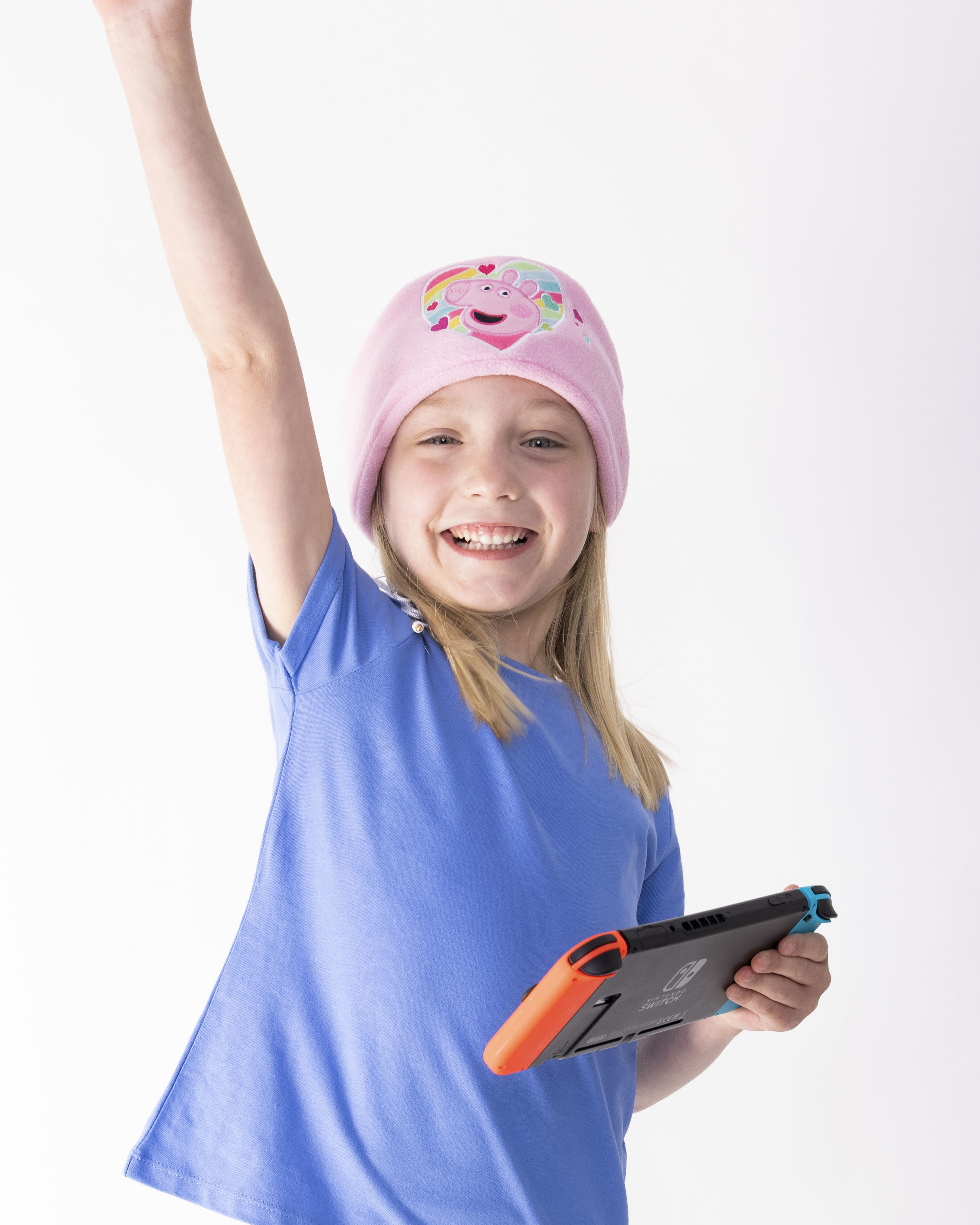 Ein junges Mädchen in einem rosa Peppa Wutz Hut und blauem Kleid, hält Nintendo Switch, lächelt.