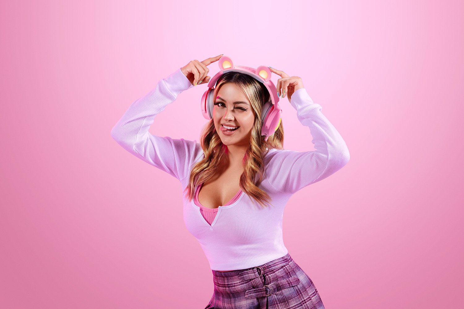 Blondynka mruga, nosząc różowe słuchawki z uszami kota, na różowym tle.