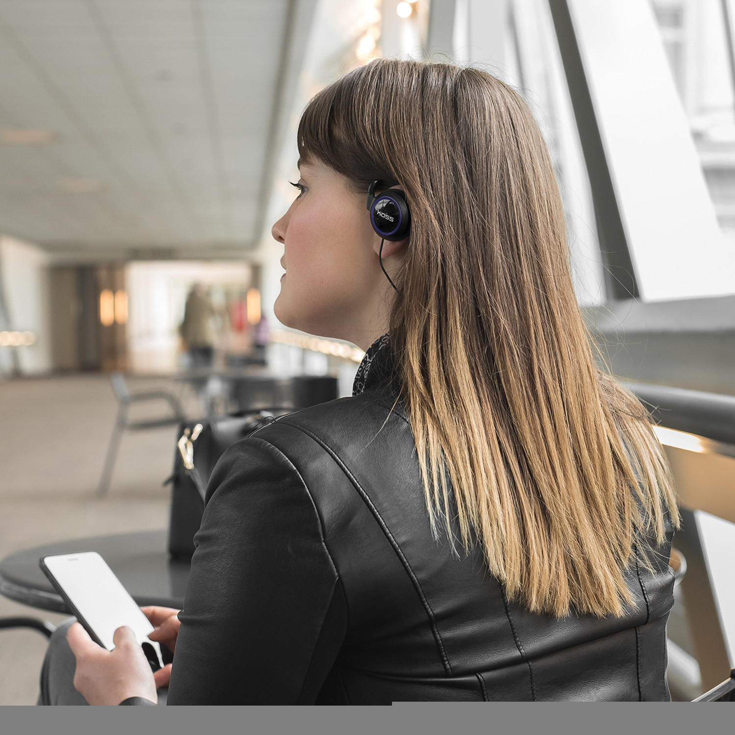 Kobieta w czarnej kurtce i słuchawkach, trzymająca telefon, w nowoczesnym budynku.