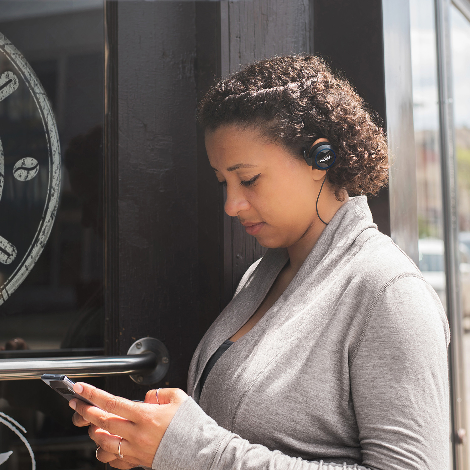 Kobieta w słuchawkach patrzy na swój telefon, stojąc przed sklepem.