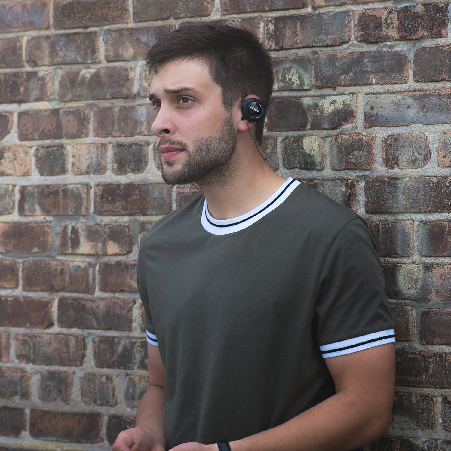 Mężczyzna w oliwkowej zielonej koszuli i czarnych słuchawkach, oparty o ceglaną ścianę na zewnątrz.