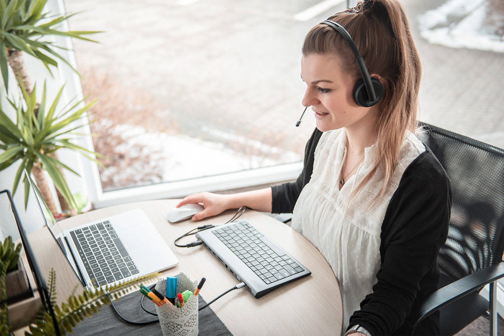 Kobieta w zestawie słuchawkowym, pracująca na laptopie przy biurku z akcesoriami i widokiem z okna.