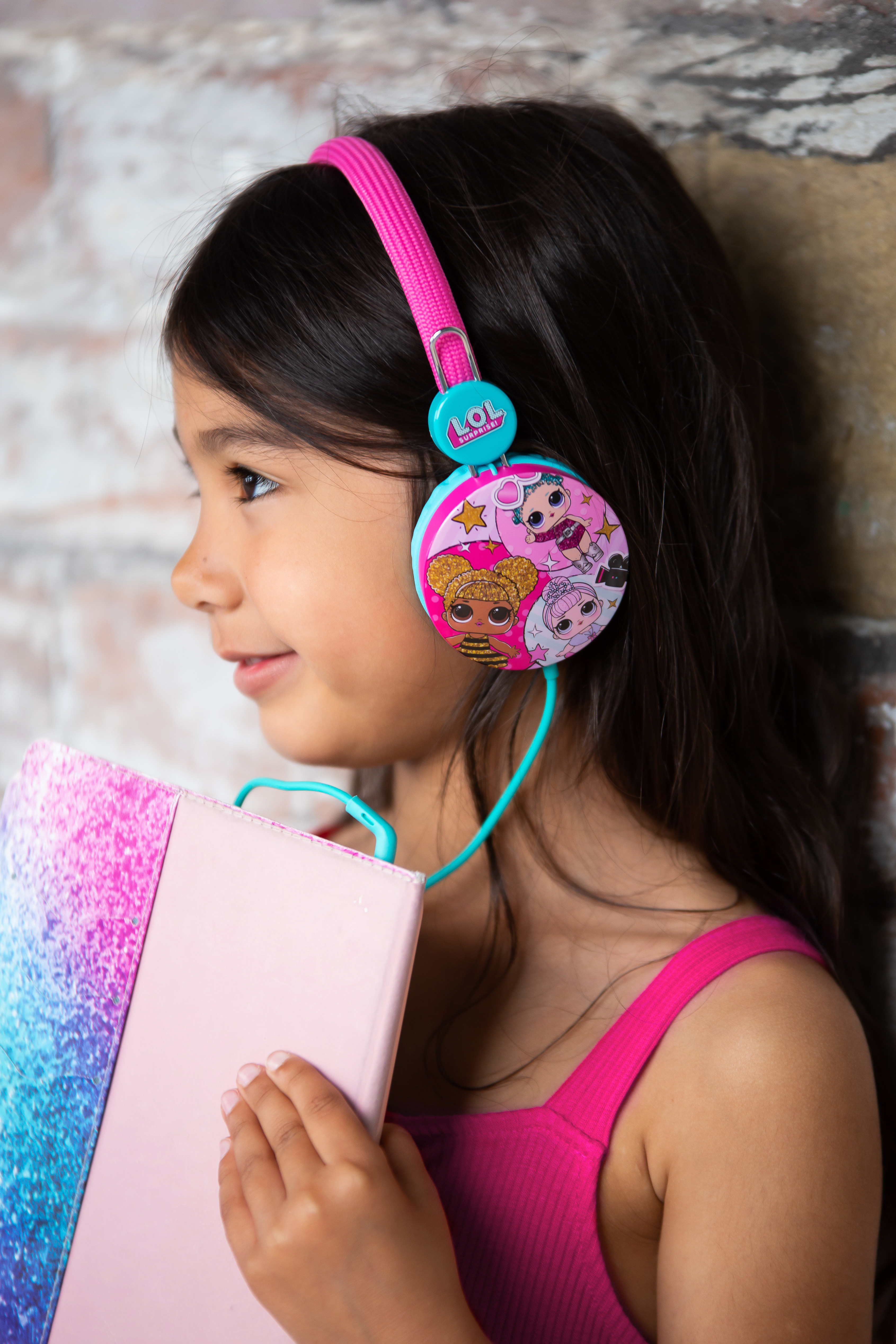 Młoda dziewczyna w różowych słuchawkach LOL trzyma książkę, opierając się o ceglany mur.