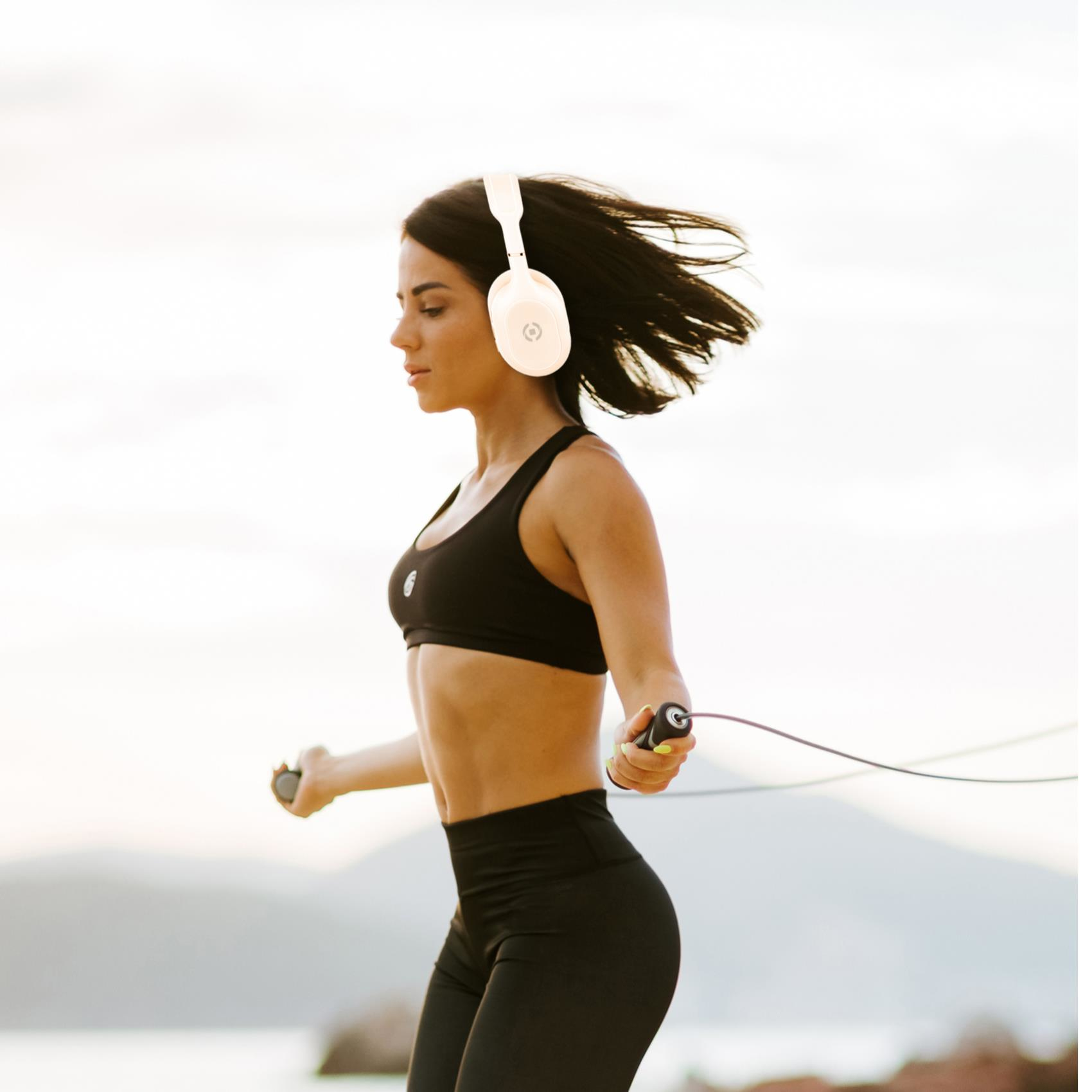 Donna che salta la corda, indossa reggiseno sportivo e leggings neri, cuffie bianche. Sfondo sfocato.