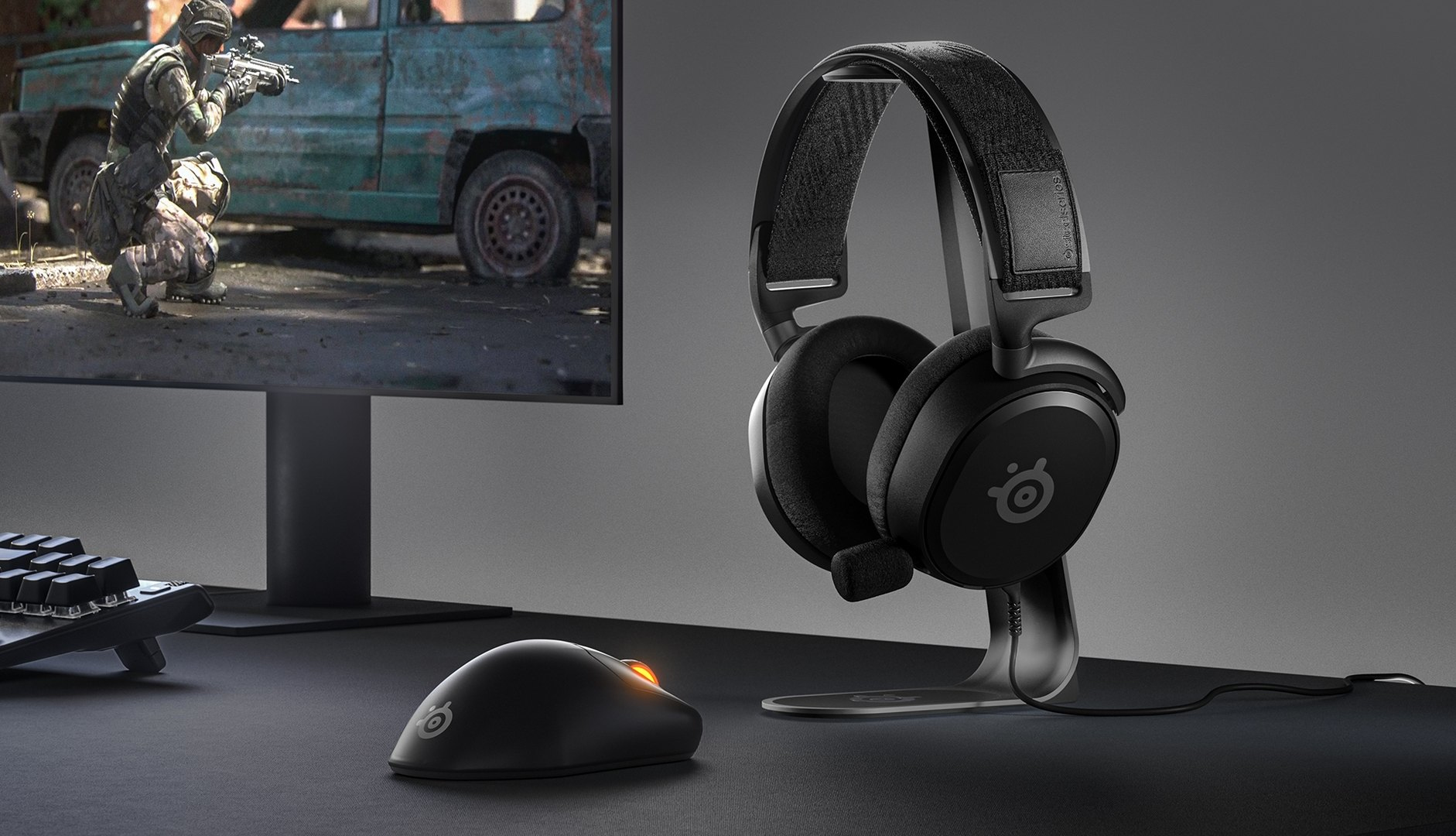 Schwarzes Gaming-Headset und Maus auf einem dunklen Schreibtisch mit Monitor und unscharfem Hintergrund.