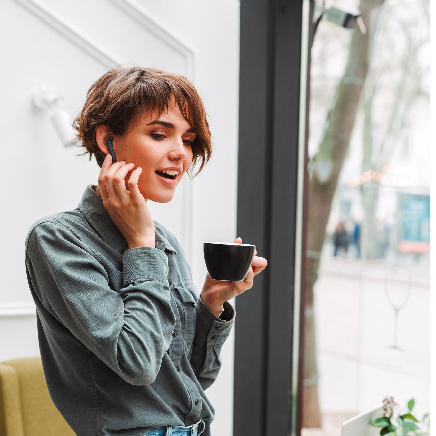 Donna con auricolari e tazza di caffè. Indossa una camicia verde, all'interno vicino a una finestra.