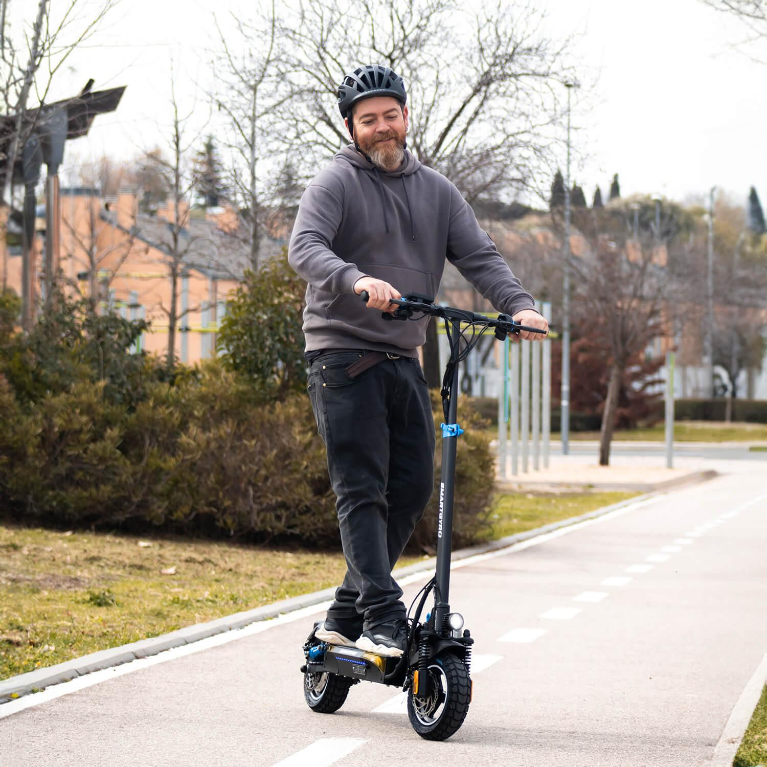 Mężczyzna jeżdżący na hulajnodze elektrycznej po utwardzonej ścieżce. Ma na sobie kask i bluzę z kapturem.