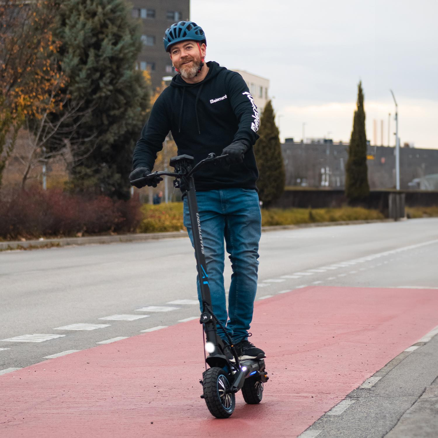 Un hombre monta un patinete eléctrico en un carril rojo. Lleva casco y guantes. Edificios al fondo.