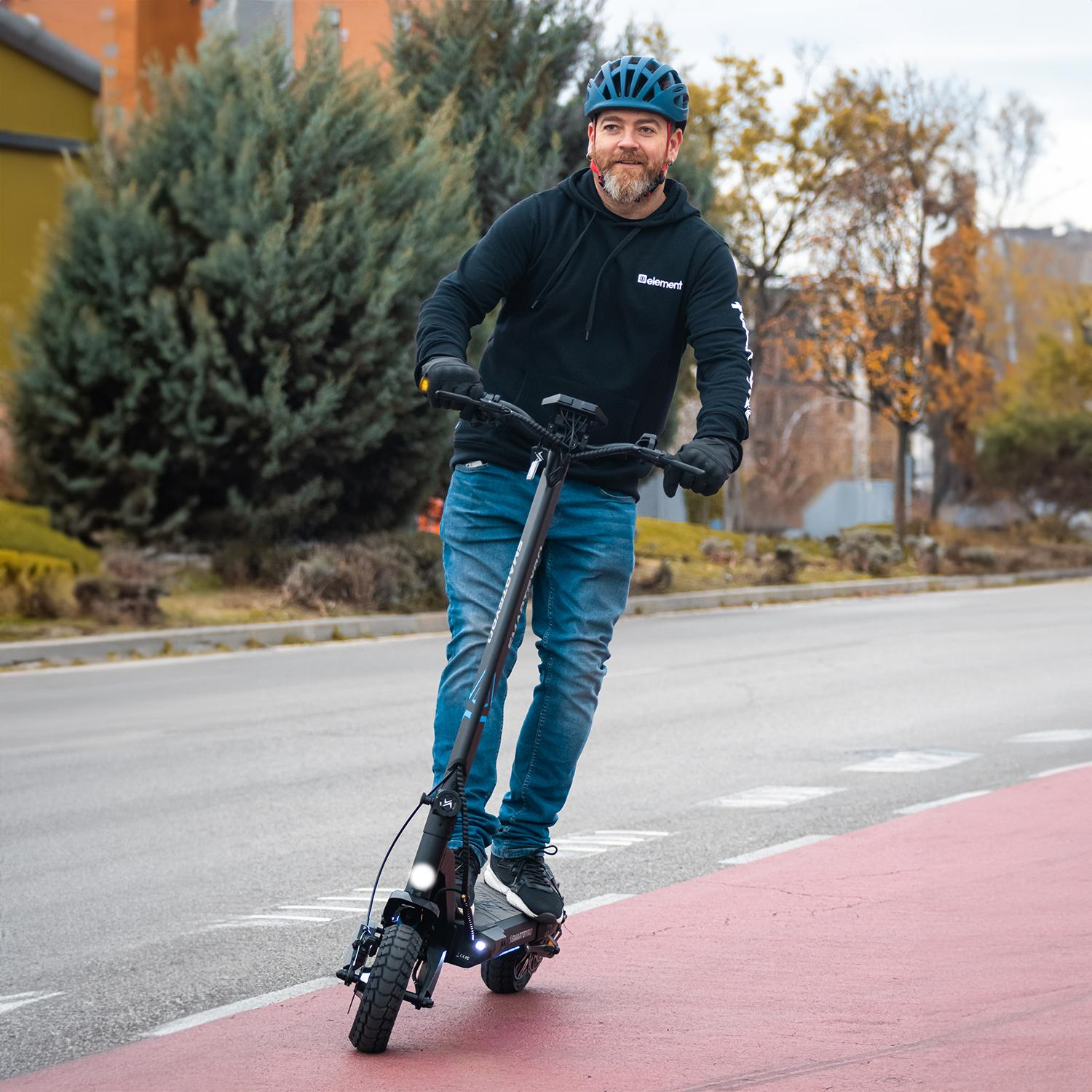 Hombre en scooter negro en carril bici rojo. Usa casco azul y jeans. Árboles al fondo.