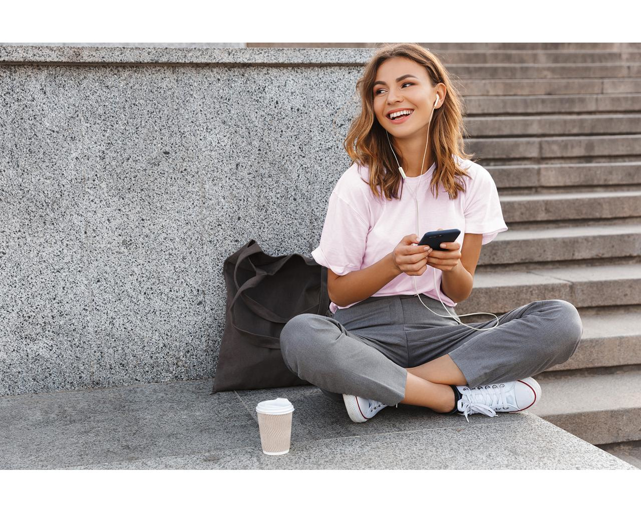 Kobieta siedząca na schodach, uśmiechnięta, w słuchawkach, trzymająca telefon.