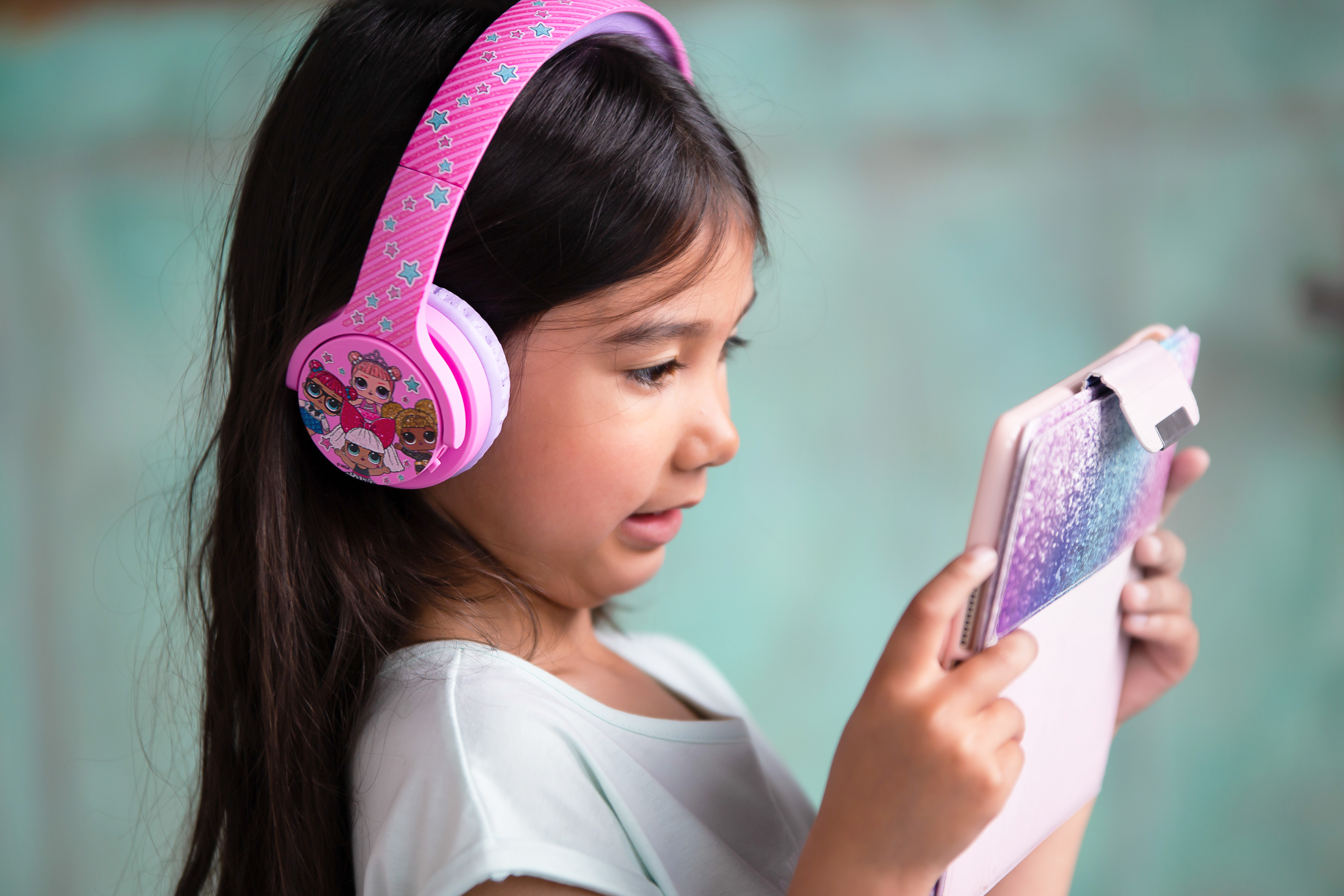Joven con auriculares rosas, mirando una tableta. El fondo es una pared turquesa.