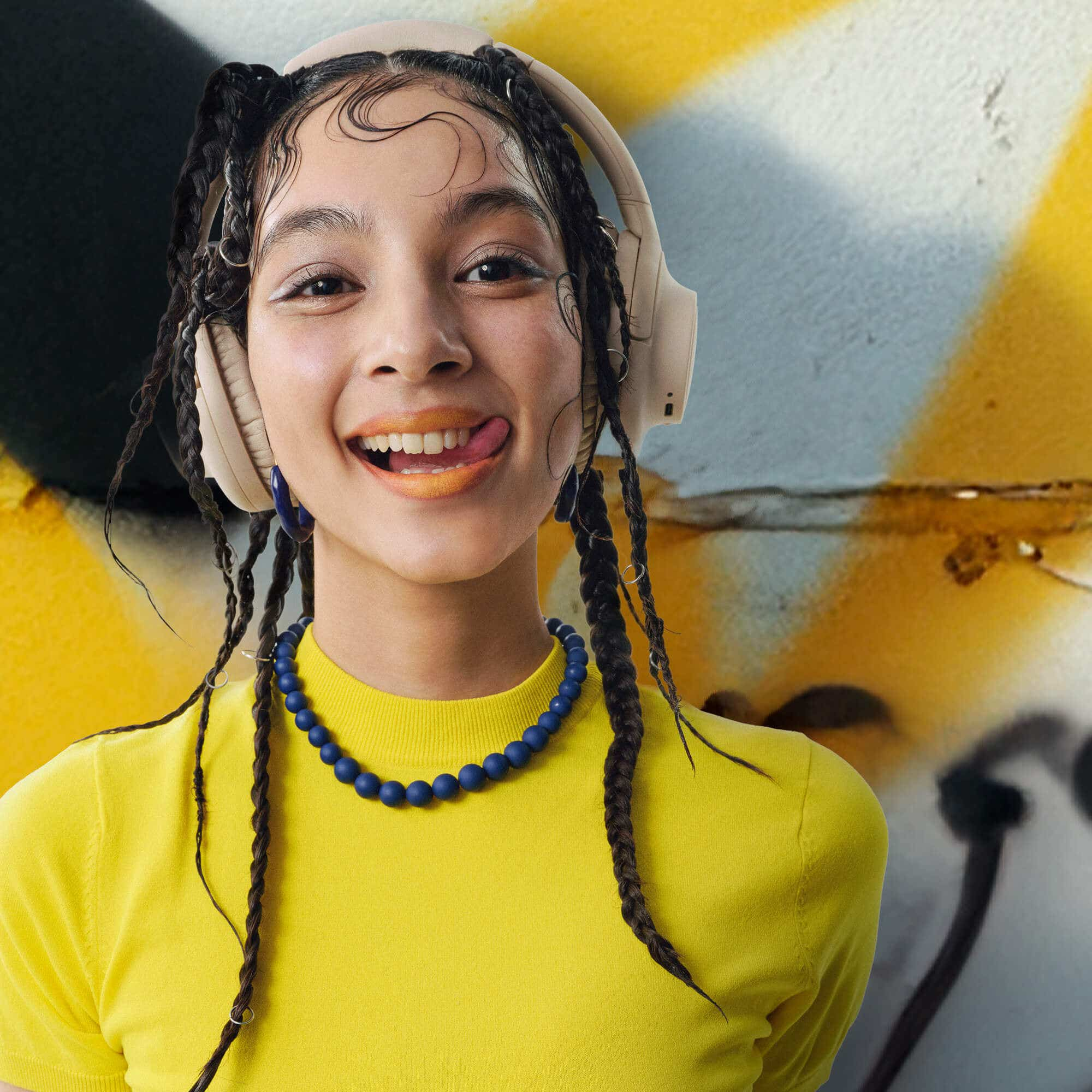 Giovane donna sorride, indossa cuffie e collana, su sfondo giallo e graffiti.