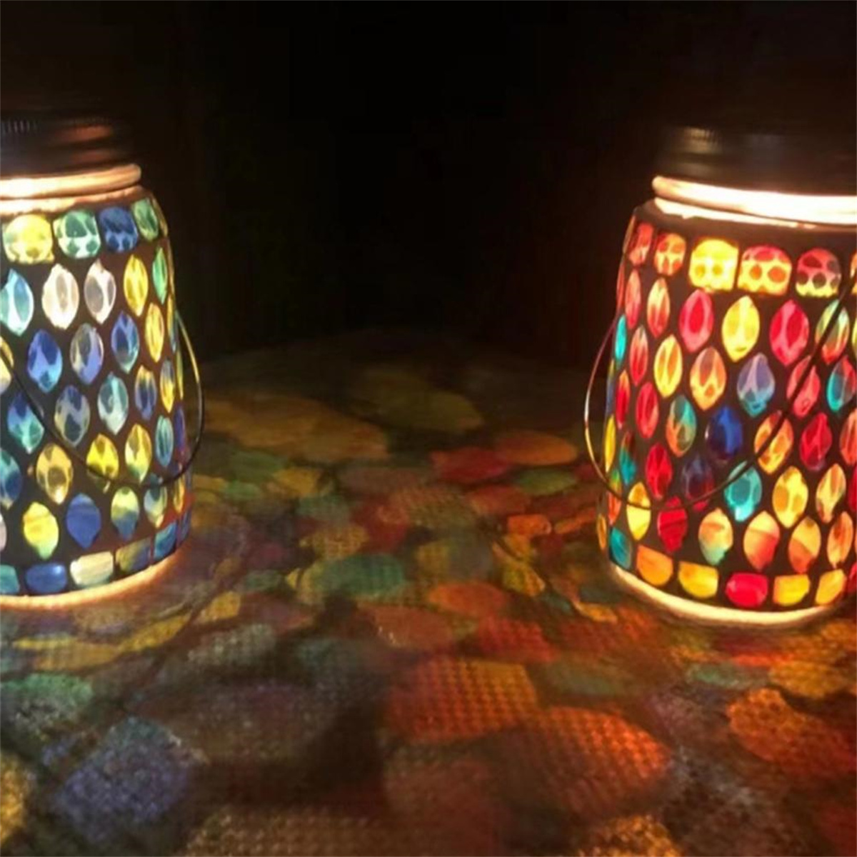 Zwei farbenfrohe Buntglasgläser, von innen beleuchtet, werfen farbenfrohe Lichtmuster auf eine dunkle Oberfläche.
