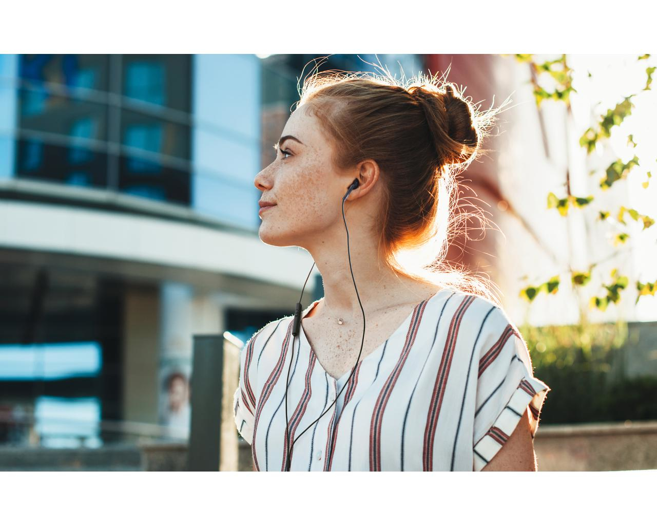 Kobieta z rudymi włosami i piegami w czarnych słuchawkach na zewnątrz. Tło jest rozmyte.