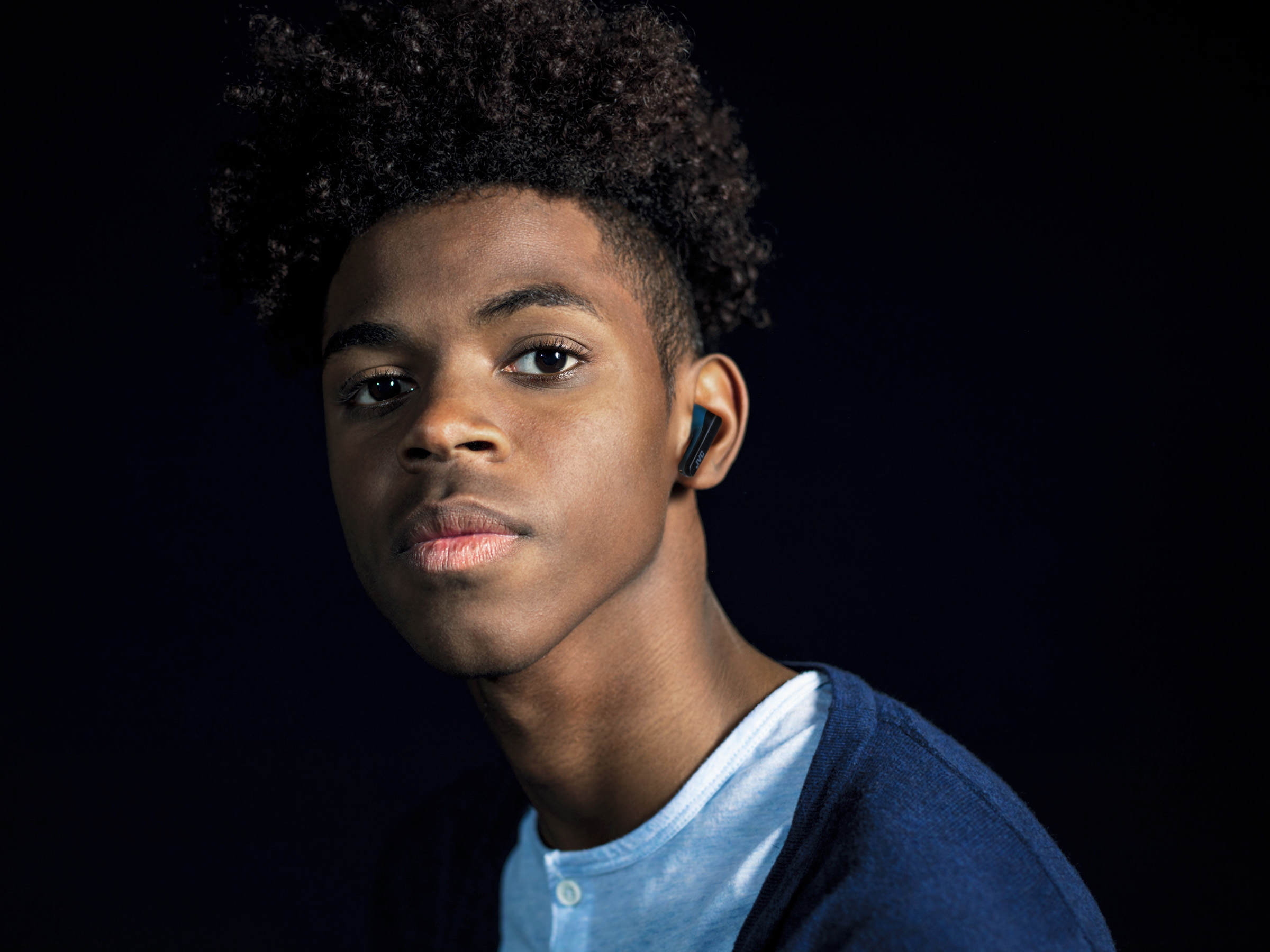 Młody czarny mężczyzna z wkładką douszną w lewym uchu, patrzący w kamerę.