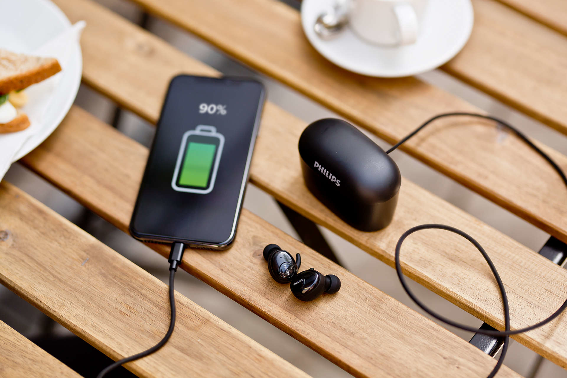 Smartfon w trakcie ładowania, obok słuchawki i etui ładujące na drewnianym stole.