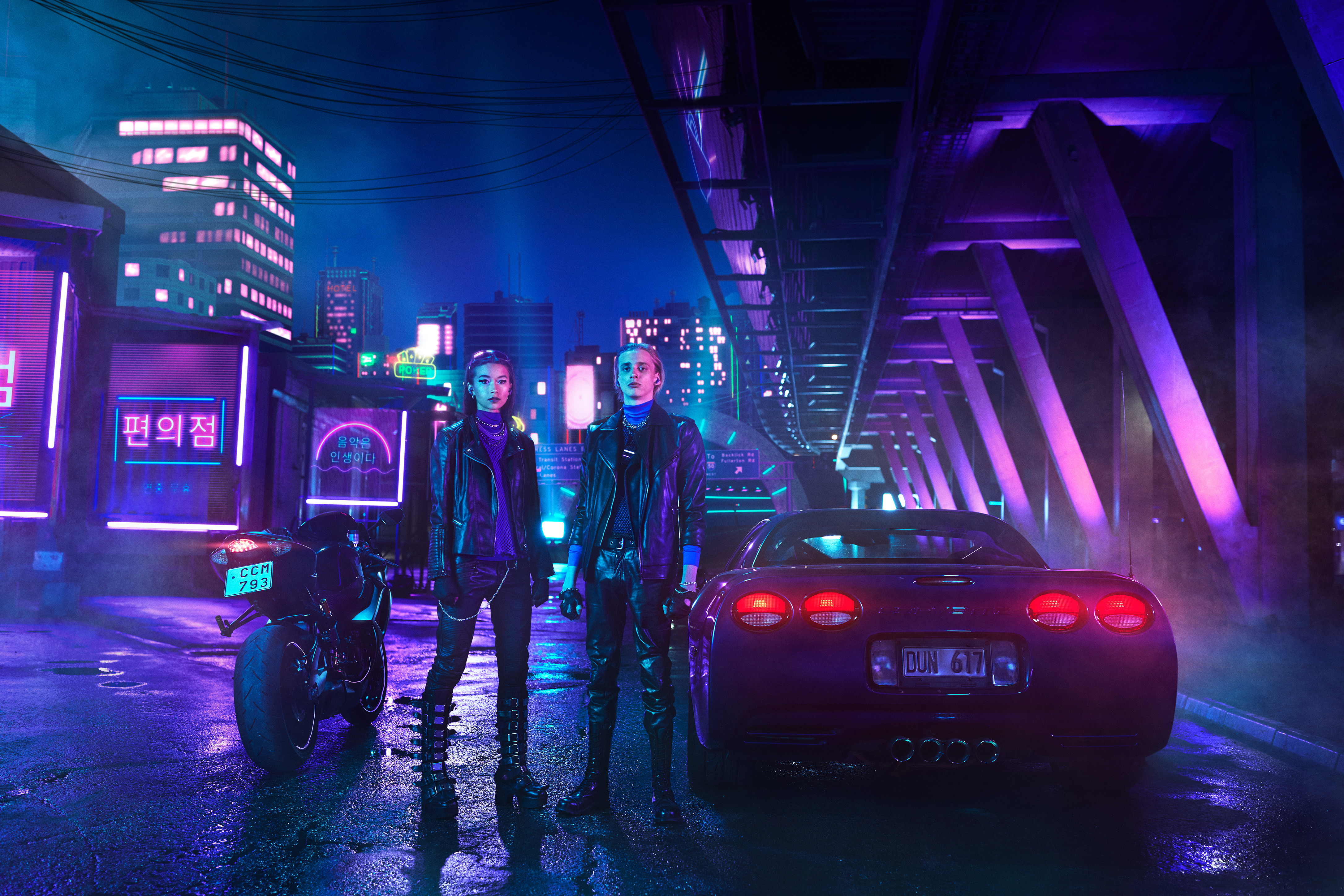 Dwie osoby stoją na oświetlonej neonami ulicy z samochodem i motocyklem. Budynki w tle.