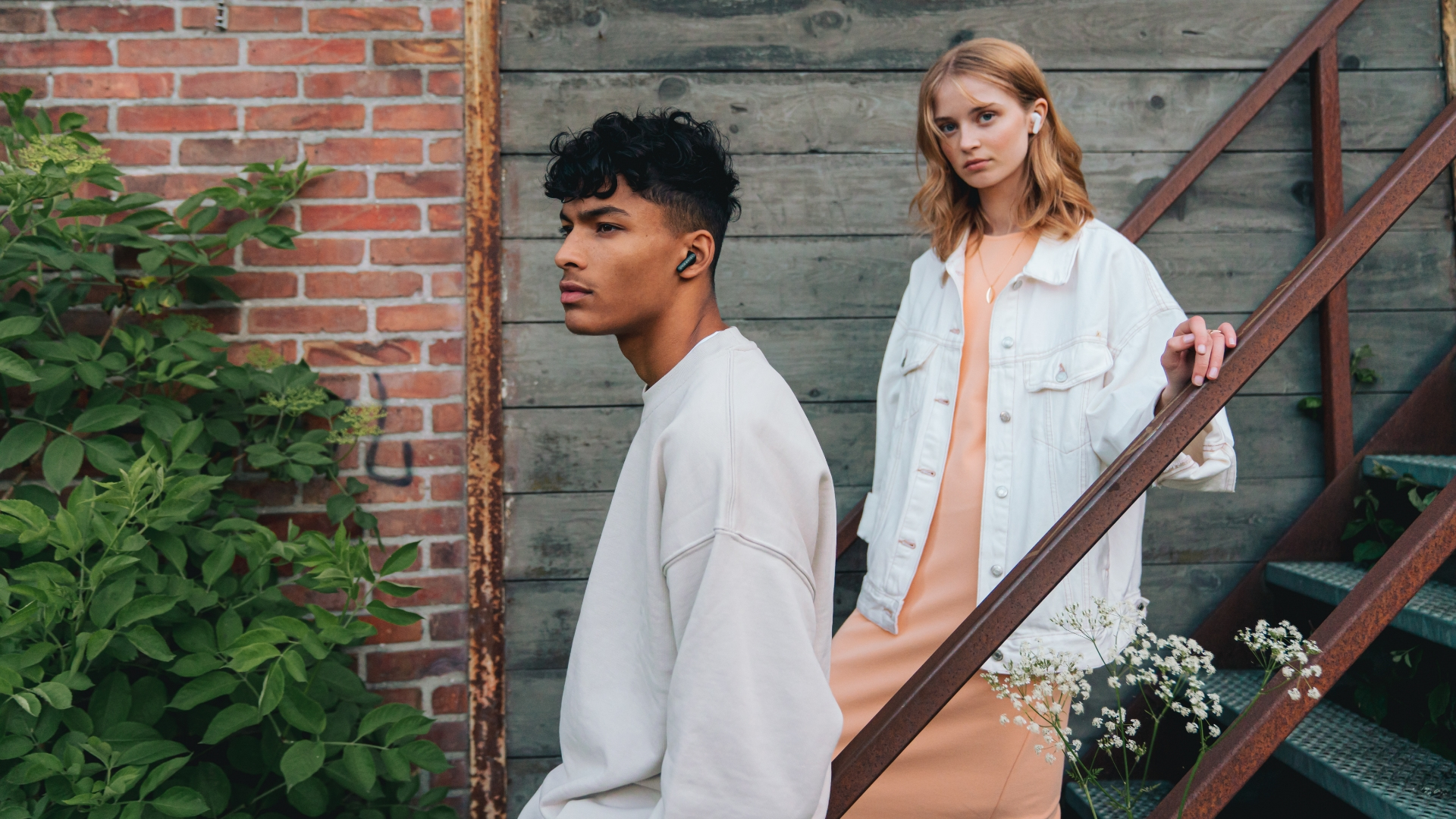 Dwie osoby, z których jedna ma na sobie słuchawki, stojące w pobliżu ceglanej ściany i metalowych schodów, na zewnątrz.